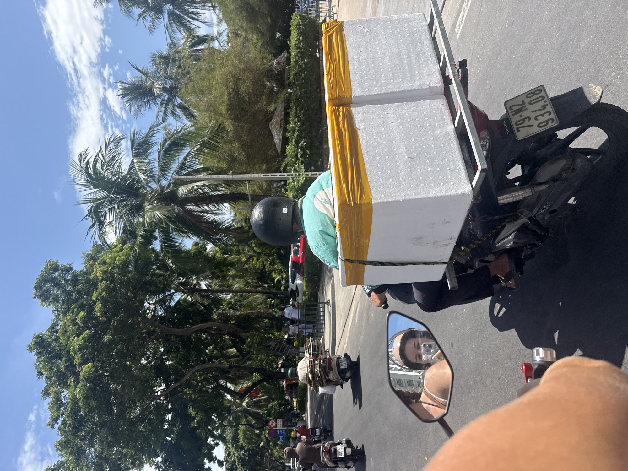 In Nha Trang, Vietnam, a man on a motorbike wearing a black helmet and a light blue shirt rides on the right side of a paved road. He is transporting two large, white styrofoam boxes secured to the rear of his motorbike with yellow tape. The boxes are stacked one behind the other on a metal rack. Behind him, a woman is reflected in the motorbike's side mirror; she is wearing a light-colored top and a helmet, and is holding a smartphone, likely taking a selfie. The road is lined with lush green trees, including palm trees, and the sky is blue with scattered white clouds. Other motorbikes and cars are visible in the background, indicating urban traffic. A street sign in Vietnamese is also present. The overall scene captures a typical mode of transportation and delivery in a bustling Vietnamese city.