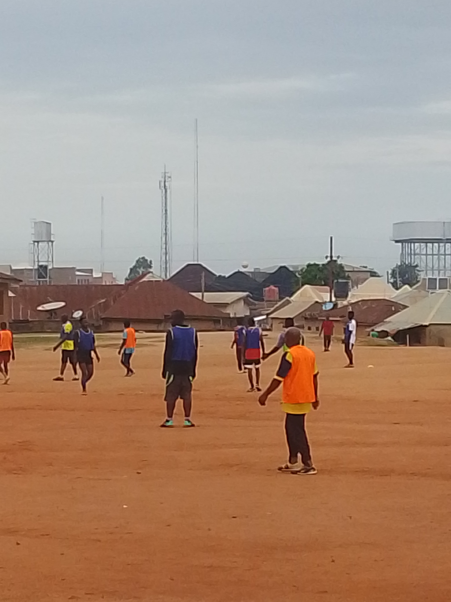 A group of people are playing a game on a dirt field in Bukuru, Nigeria. The players are wearing various colored vests, including orange and blue. One person is throwing a disc in the air. In the background, there are buildings, including a water tower and telecommunication towers. The sky is overcast.