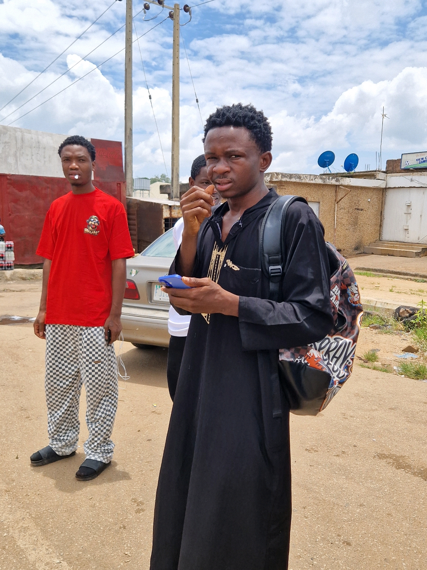 In Jos, Nigeria, a young man wearing a red t-shirt with a graphic print and checkered pants stands to the left. He has a piece of white material in his mouth. To his right, another young man in a black garment and a backpack with colorful graffiti is holding a blue mobile phone. A third person, partially visible, is standing behind them. The background shows a street with a car, buildings, and overhead utility lines against a cloudy sky.