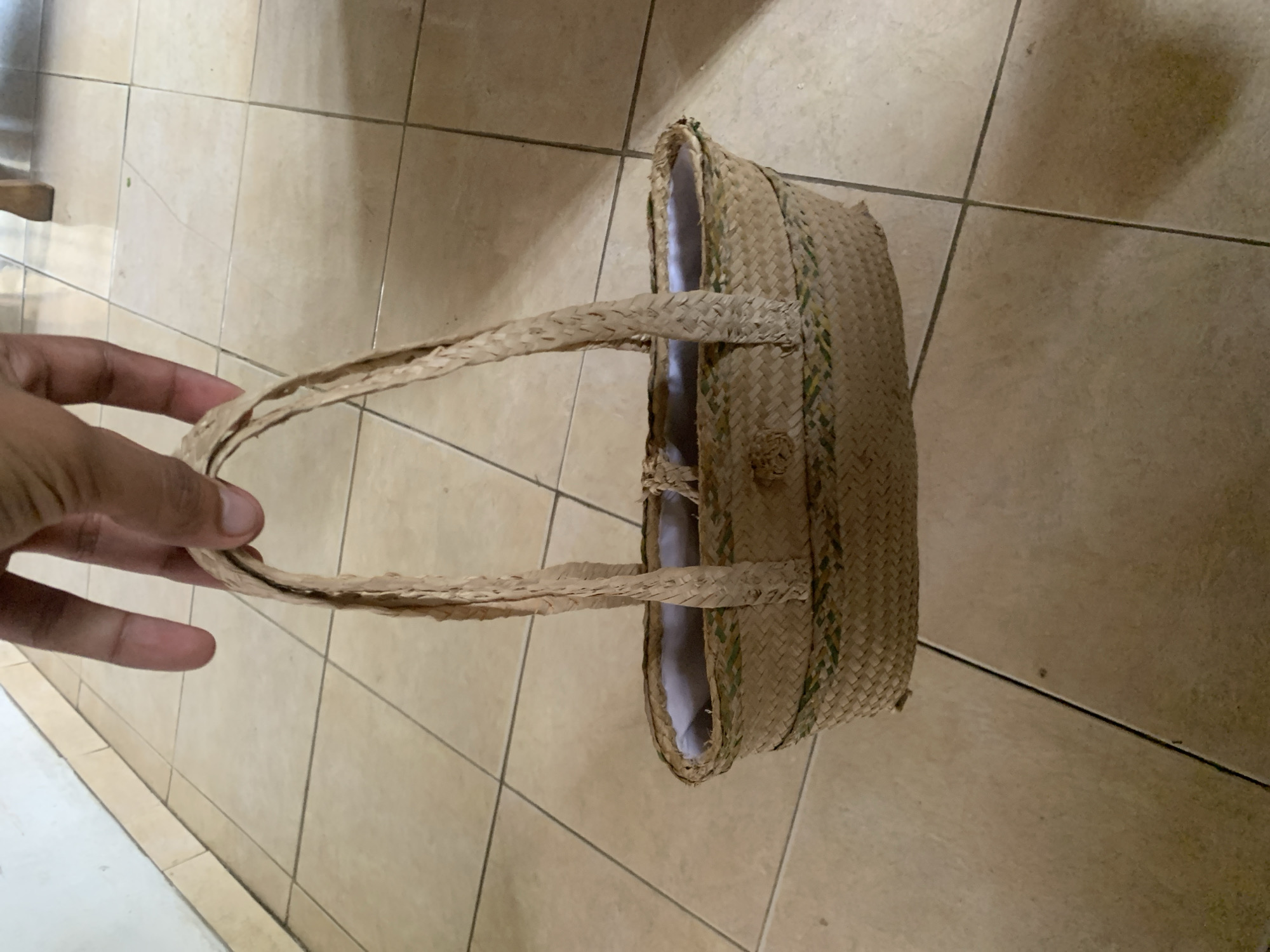 The image, rotated 90 degrees clockwise, displays a person's left hand holding a woven straw or wicker bag against a tiled floor background. The hand is visible on the left side of the frame, gripping one of the bag's two braided handles. The bag, positioned towards the right side, is crafted from natural, light brown woven material, possibly raffia or palm leaves, with faint greenish strands subtly integrated into its weave. A light-colored fabric lining is visible inside the bag's opening. The background consists of large, square, beige or cream-colored ceramic floor tiles with dark grey grout lines, suggesting an indoor or covered outdoor setting. The activity shown is simply a hand holding the handcrafted bag. There are no specific time of day or weather cues, and no emotions are discernible. No text is visible in the image. The scene depicts a simple, everyday object in Santa Ana, Venezuela.