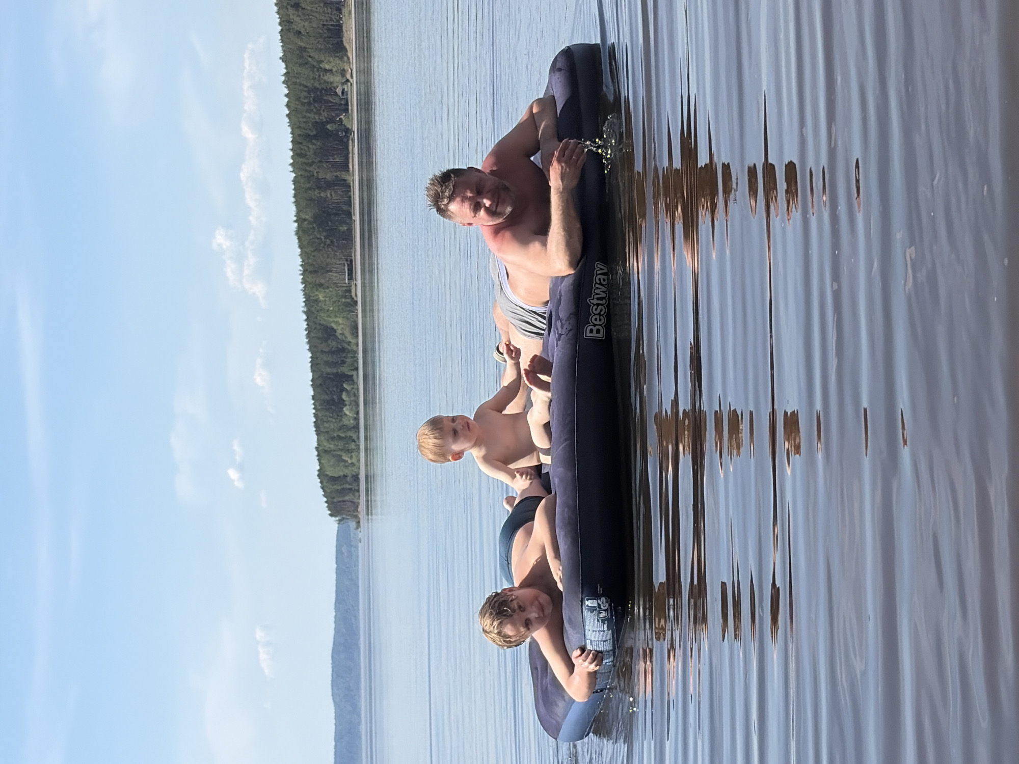 The image shows three people on an inflatable raft in a body of water, likely a lake, surrounded by trees. From top to bottom, the individuals appear to be a man, a young child, and an older child or young teen. The inflatable raft is dark blue/black with the logo "Bestway" visible. The background shows a forested shoreline, a light blue sky, and possibly some clouds. The image suggests a summer day setting. The location is identified as Vekoshinka, Russia.