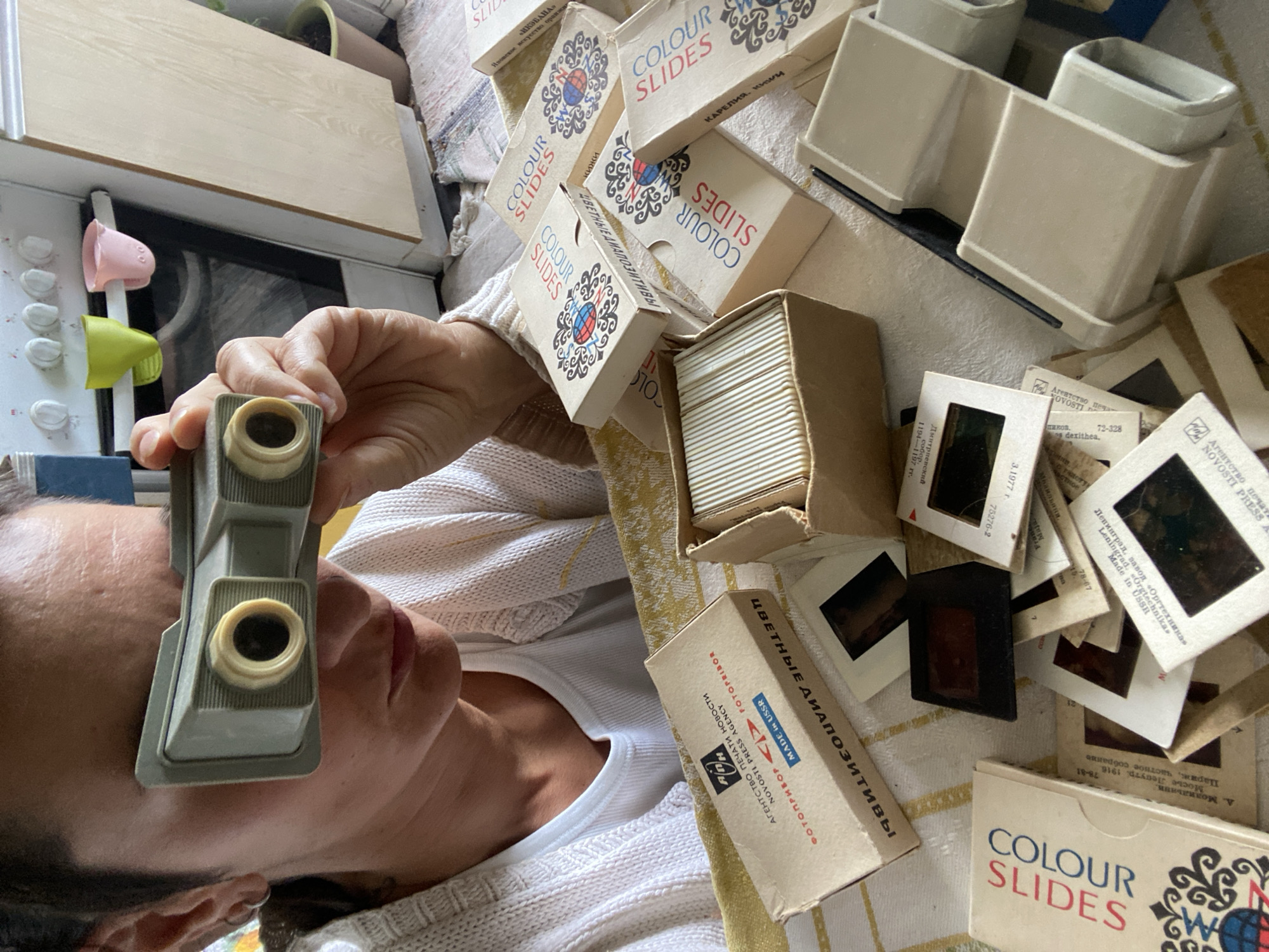 In Moscow, Russia, a person lies down, intently engaged in viewing old photographic slides through a handheld, vintage beige-grey slide viewer. The scene is indoors, likely a home, with a microwave oven and a wooden cabinet visible in the background, suggesting a kitchen or living area.
The person is surrounded by numerous cardboard boxes and loose mounted slides, implying a collection or a session of reminiscing. Many of the boxes are labeled "COLOUR SLIDES" in English, accompanied by Cyrillic script. Other boxes and individual slide mounts feature prominent Russian text.
Notable details from the visible text include "ЦВЕТНЫЕ ДИАПОЗИТИВЫ" (Colour Slides in Russian) and the recurring phrase "MADE IN USSR," indicating the origin of these photographic materials. Several boxes carry the name "АГЕНТСТВО ПЕЧАТИ НОВОСТИ" (Novosti Press Agency), with "NOVOSTI PRESS AGENCY" also in English, and "ФОТОПРИБОР" (Fotopribor). Specific manufacturing details like "Ленинград, завод «Оргтехника»" (Leningrad, Orgtechnika plant) are also present. Themes for slide sets are visible, such as "«ИКЭБАНА» Ивовское искусство оранжи" (Ikebana, Japanese art of arrangement) and "КАРЕЛИЯ, кими" (Karelia, Kemi), suggesting travel or cultural topics. There are also references to artists or subjects like "А. Модильяни" and "Мосье Лепутр. 1916," along with dates like "3.1977 г." and other catalog numbers (e.g., 72-328, 75276-2). The light suggests daytime. The activity evokes a sense of nostalgia and focused concentration.