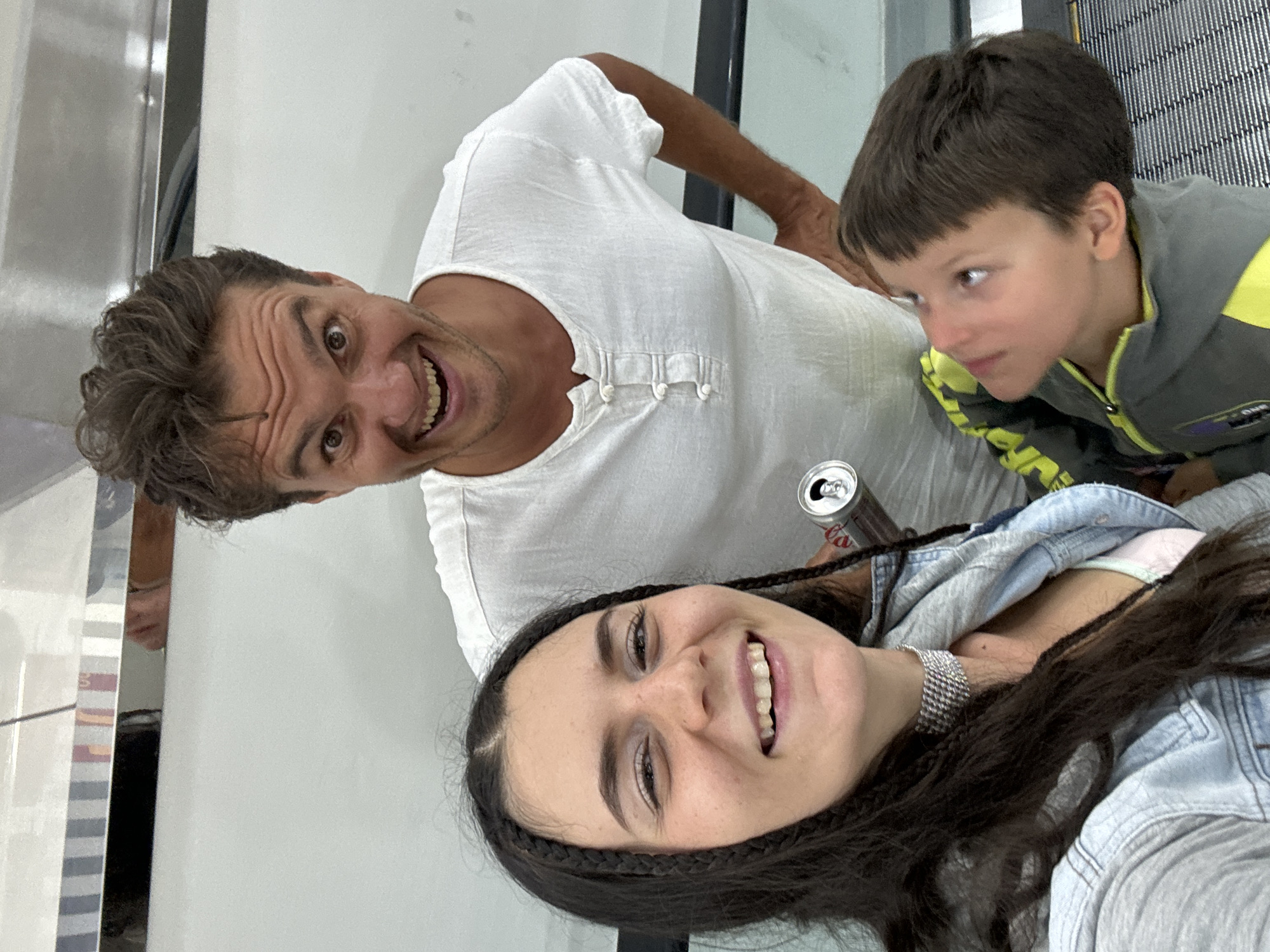 A candid, off-center shot captures a trio of individuals. A man with dark hair, wearing a white shirt with distinctive toggle closures at the neck, grins directly at the camera. To his right and slightly above him is a younger boy with short brown hair, who glances off to the side with a serious expression. At the bottom of the frame, a woman with dark hair, partially braided, smiles, wearing a denim jacket and a silver necklace. A can of Coca-Cola rests near her head. The background consists of a light grey wall and a grated metal surface, suggestive of an escalator or elevator. There is no clear indication of the location being Phu Thuy Ward, Vietnam. The scene depicts a casual moment, seemingly taken during travel or in a public space.