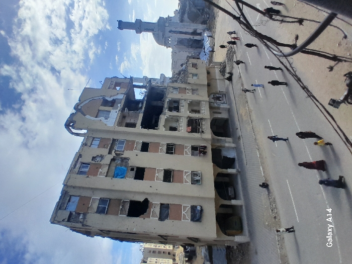 The image depicts a street scene in Khan Yunis, Palestinian Territory, characterized by significant structural damage. A multi-story, light-colored building, approximately seven to eight floors tall, dominates the left-center, exhibiting severe destruction to its upper floors and right facade. Numerous windows are broken or missing, and sections of the exterior wall are collapsed, revealing interior structural elements. Small impact marks are visible across its surface, and a blue tarp hangs from a window on the third floor. To the right and behind this building, a mosque with a tall minaret and a dome is visible, also showing signs of damage, particularly around its dome and upper minaret.
The foreground features an asphalt road with white lane markings. Approximately 20 individuals, including adults and at least two children, are dispersed across the road and sidewalks. Most are walking, generally clad in dark clothing; one individual wears red, and a child wears yellow. One person is seated on the ground near the base of the damaged building. To the far right, partially obscured utility poles and numerous dark wires cross the frame. A motorcycle or scooter is partially visible in the lower right foreground. Beyond the main damaged building, additional structures in varying states of destruction are visible under a partly cloudy sky. A vehicle is parked under the ground-level arches of the heavily damaged building.