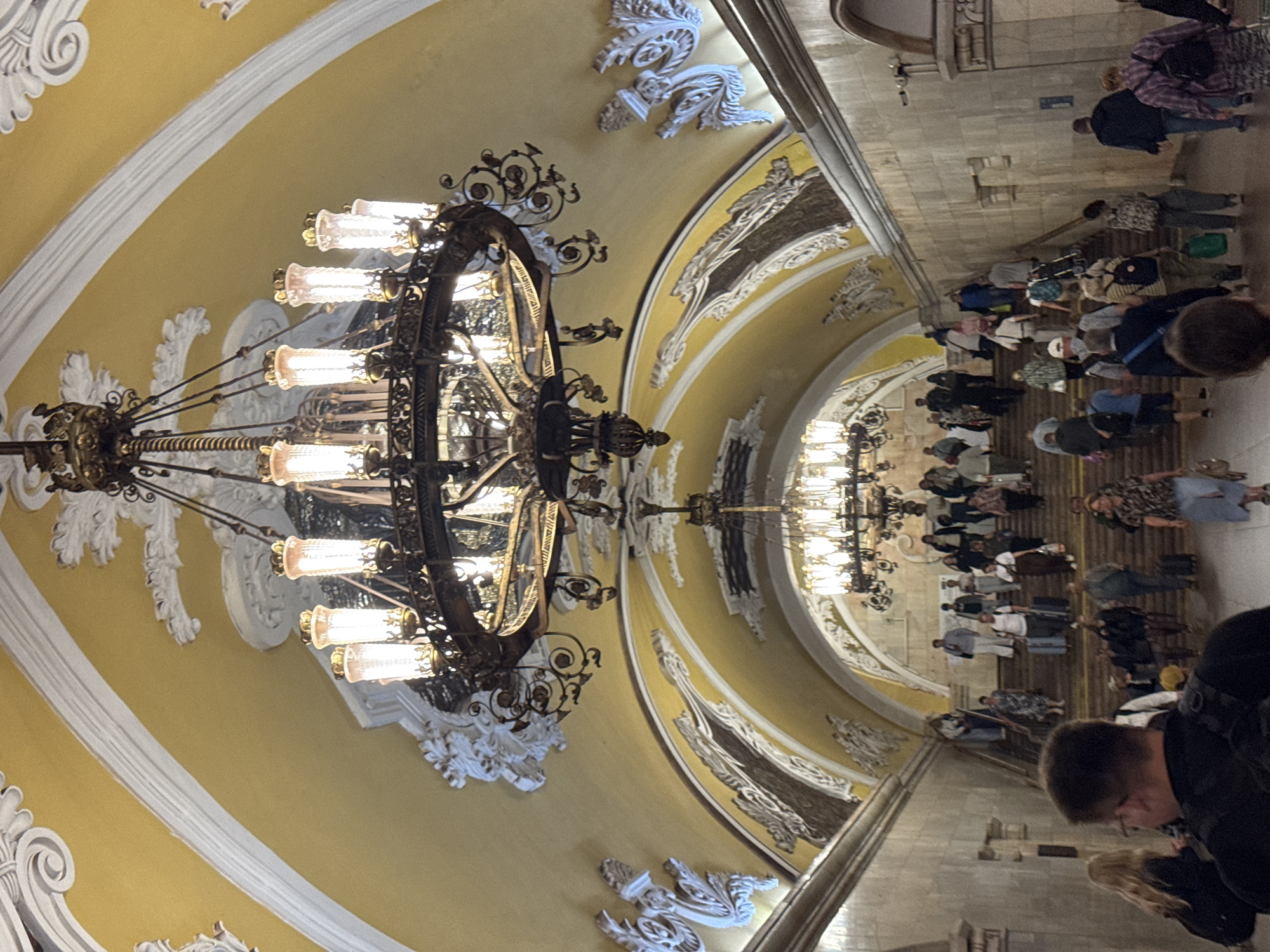 The image depicts a grand staircase within a Moscow Metro station, likely Komsomolskaya Station due to its ornate architectural style. The station features a high, arched ceiling painted a vibrant yellow, adorned with elaborate white stucco moldings. Two large, multi-tiered chandeliers, one in the foreground and another further down the stairs, illuminate the scene with warm light. The chandeliers are intricate metal structures with numerous glass lamps.
The staircase itself is made of stone, with wide steps and a substantial stone balustrade. A crowd of people are seen ascending and descending the stairs, many carrying luggage, suggesting it's a busy travel hub. The individuals are diverse in age and attire, dressed in casual clothing. Some are walking alone, while others are in small groups. The overall impression is one of grandeur and public transit activity within a richly decorated subway environment.