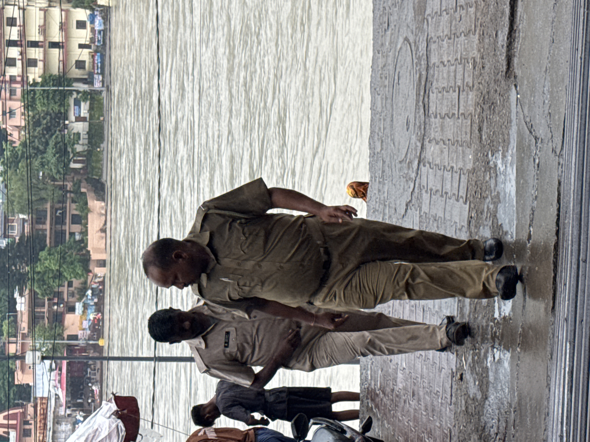 Two men, likely police officers given their uniforms, walk down a wet, patterned walkway next to a wide, murky body of water, possibly the Ganges River. The man on the right is in the foreground, facing away from the camera and looking to his left. He wears a khaki uniform with a short-sleeved shirt and pants. His companion, to his left and slightly behind, is also in uniform and facing slightly away from the camera. In the background, across the water, are multi-story buildings and lush green trees, indicative of an urban area. A partially visible motorcycle is parked to the far left of the frame. The walkway appears to be outdoors, with evidence of recent rain or high water levels.