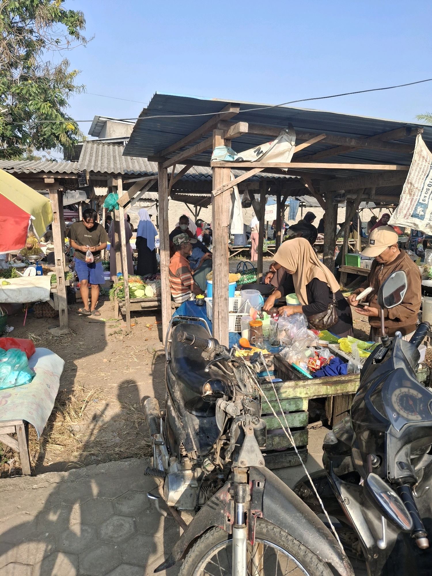 This is a street-level, outdoor, full shot of a market scene in Bulurejo, Indonesia. The market is open-air with several stalls constructed from wood and corrugated metal roofing. Sunlight casts strong shadows across the dusty ground.
In the foreground, two motorcycles are parked. The one on the left is partially disassembled, with its front wheel and fork prominently visible. The one on the right is mostly intact, facing away from the camera.
The market stalls are filled with various goods. Several individuals are present, engaged in transactions and other activities.
To the left, a stall is adorned with a red and yellow umbrella. A woman is visible behind a table with scales and produce.
Further back, a young man wearing a dark t-shirt and blue shorts stands in the middle of the market path, holding a small plastic bag.
Several other people are scattered throughout the market, some are partially obscured by the stalls and other individuals. One stall appears to be selling fresh produce, with piles of vegetables like carrots and cabbage displayed on wooden shelves. A woman wearing a beige hijab is interacting with a man wearing a brown jacket and a tan baseball cap at another stall. She is handling items on a table covered with plastic sheeting, and he is holding a piece of corn. Bags of produce and other items are visible on the table as well.
Behind the stalls, more market structures and some buildings are visible under a clear blue sky. The overall impression is of a bustling local market on a sunny day.