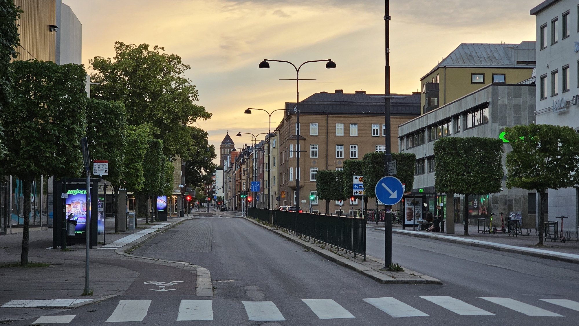 This media file depicts a street scene in Linköping, Sweden, during the early evening or dusk. The sky is filled with warm, soft hues of orange and yellow, indicating the setting sun. The weather appears calm and clear.
The scene is a street with a pedestrian crossing in the foreground. On the left side of the street, there are trees lining the sidewalk, and several illuminated digital advertising displays are visible. A signpost with bus route information is also present. On the right side of the street, there are several buildings of varying architectural styles and heights, including a modern-looking grey building with a row of awning-covered windows, and a yellow building with a grey roof. Further down the street, a line of older, multi-story buildings with colorful facades can be seen, and in the distance, a tall, dark tower with a peaked roof is visible.
The street itself is paved, with lane markings and a bicycle lane clearly delineated. A black metal fence runs along the right side of the road, separating the sidewalk from a lower level. Several streetlights with a distinctive design are mounted on poles. Small trees are planted along the sidewalk on the right side of the street, some of which are meticulously trimmed into square shapes. In the background, a few cars are visible on the road, and some people are seen sitting near a bus stop. Various objects, such as bicycles and electric scooters, are parked near the buildings.
Notable details include the traffic lights, some of which are red, suggesting that traffic is momentarily stopped. The overall mood is serene and peaceful, characteristic of a quiet city street at the end of the day.
Visible text includes "HAJPER" on one of the advertising displays, and what appears to be route numbers and letters on the bus stop sign. A blue circular sign with a white arrow pointing downwards to the right indicates a traffic direction.