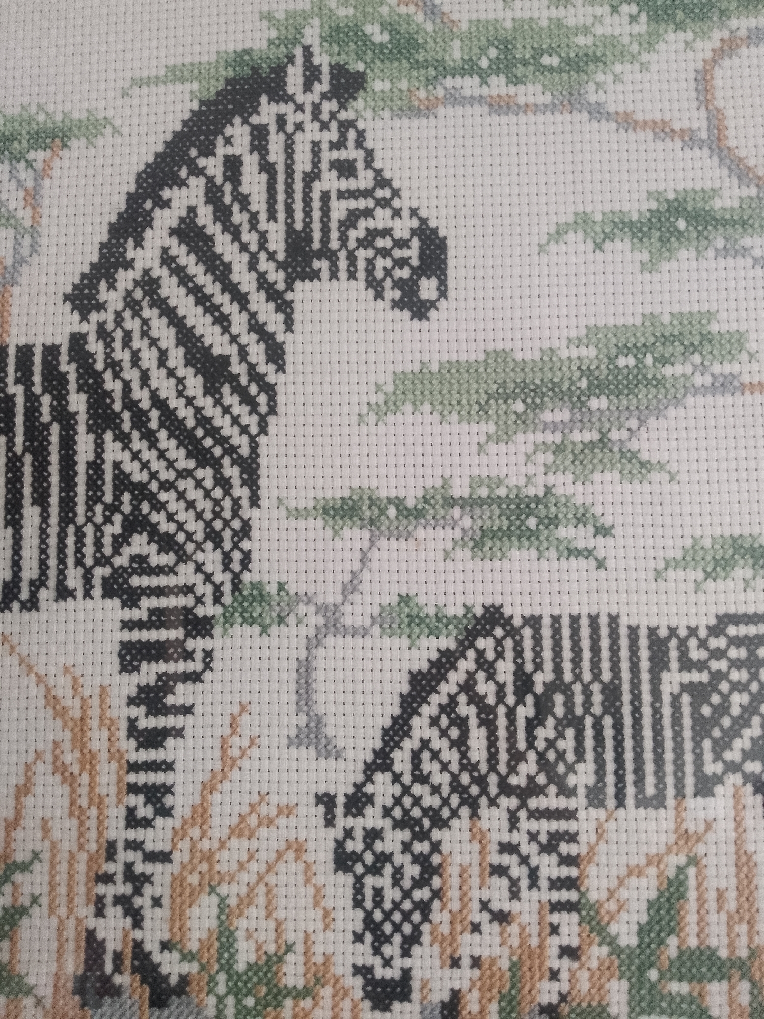 The image shows a close-up of a cross-stitch artwork featuring two zebras in a savanna-like environment. The zebras are depicted with black and white stripes, with the stitching creating a pixelated effect. The background includes stitched elements representing trees with green foliage and dry grass in shades of brown and green. The fabric used for the cross-stitch is a light-colored Aida cloth. There is no information in the image to determine the location of the image as Davao City, Philippines.
