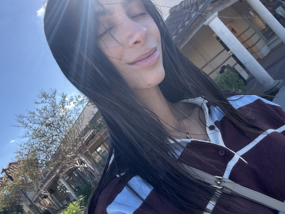 A woman with long, dark hair is facing the sun with her eyes closed. She wears a burgundy and light blue striped cardigan and has two necklaces. In the background, there is a building with columns, trees, and a blue sky. The sunlight casts a bright glare on the image.