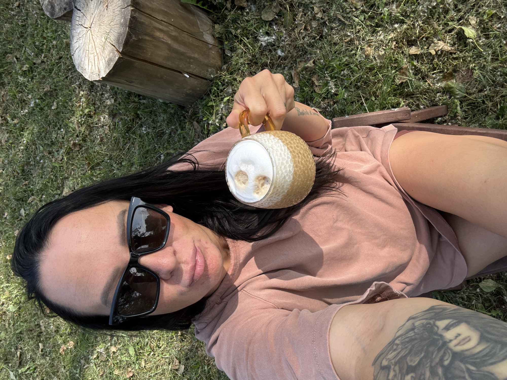 A woman with dark hair and sunglasses is holding a mug of coffee outdoors. She is wearing a pink shirt and has a tattoo on her arm. The background shows grass, leaves, and a wooden log. The scene appears to be a park or backyard. The image does not contain enough information to determine a specific location within Chaikovsky, Russia.