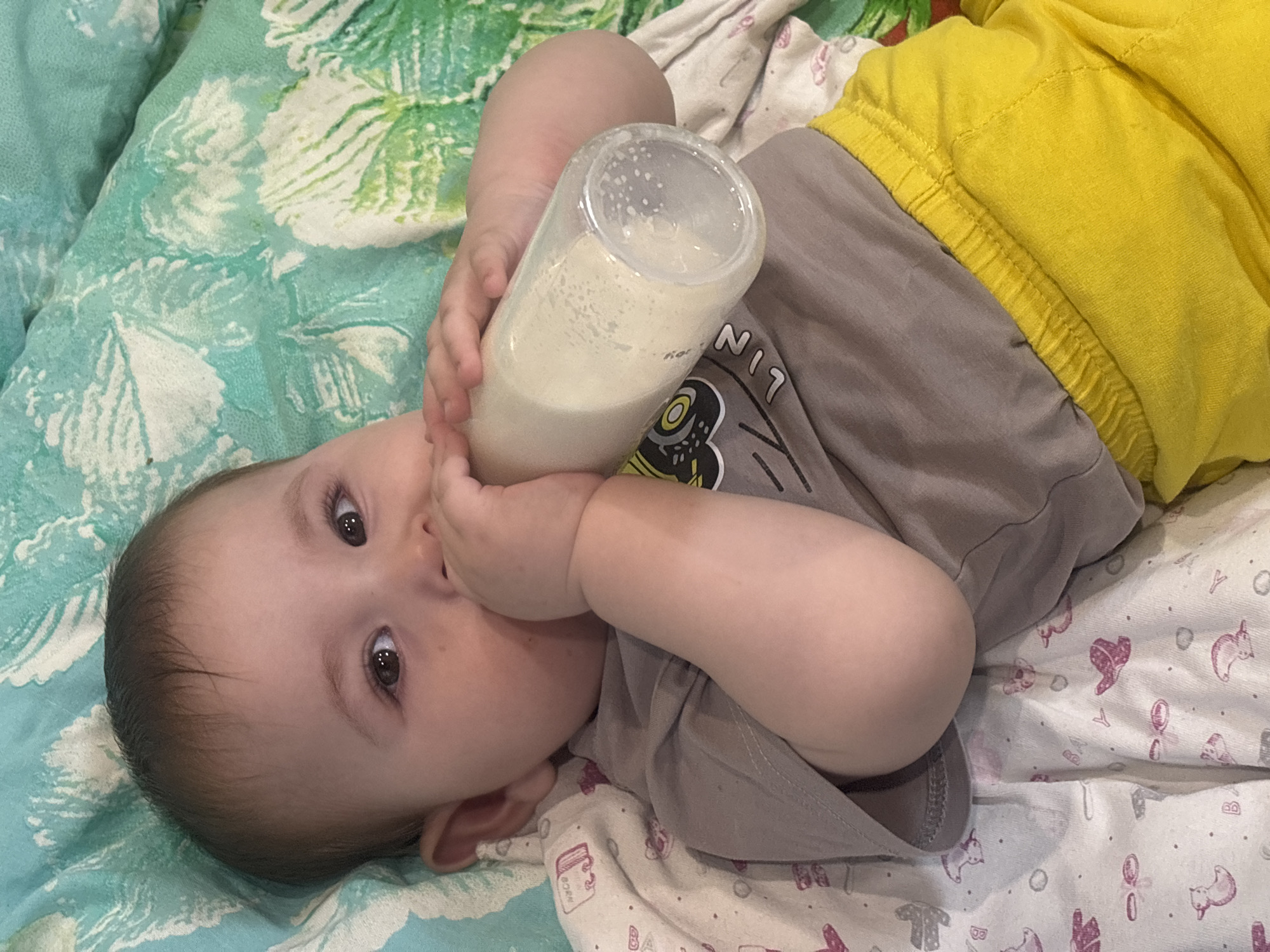 The scene depicts an infant lying on its back in an indoor setting within the city of Chaikovsky, Russia. The infant, with dark hair and eyes, is positioned centrally, facing slightly towards the viewer's right. It is wearing a light gray short-sleeved top with a partially visible graphic featuring a yellow circle and black outlines, and bright yellow elastic-waisted shorts. Both of the infant's hands grasp a clear plastic baby bottle, which contains a white liquid, likely milk or formula, exhibiting air bubbles. The infant's mouth is engaged with the nipple of the bottle.
The infant rests on two distinct layered fabrics. Beneath its head and upper torso is a teal or light green blanket or sheet with a faded white, organic, possibly leaf or abstract floral pattern. Underneath the infant's lower body and right arm is a white blanket or sheet patterned with small, repeating pink and purple illustrations of various objects including ducks, bows, hats, and letters such as 'B', 'Y', and 'N_b_c_'.