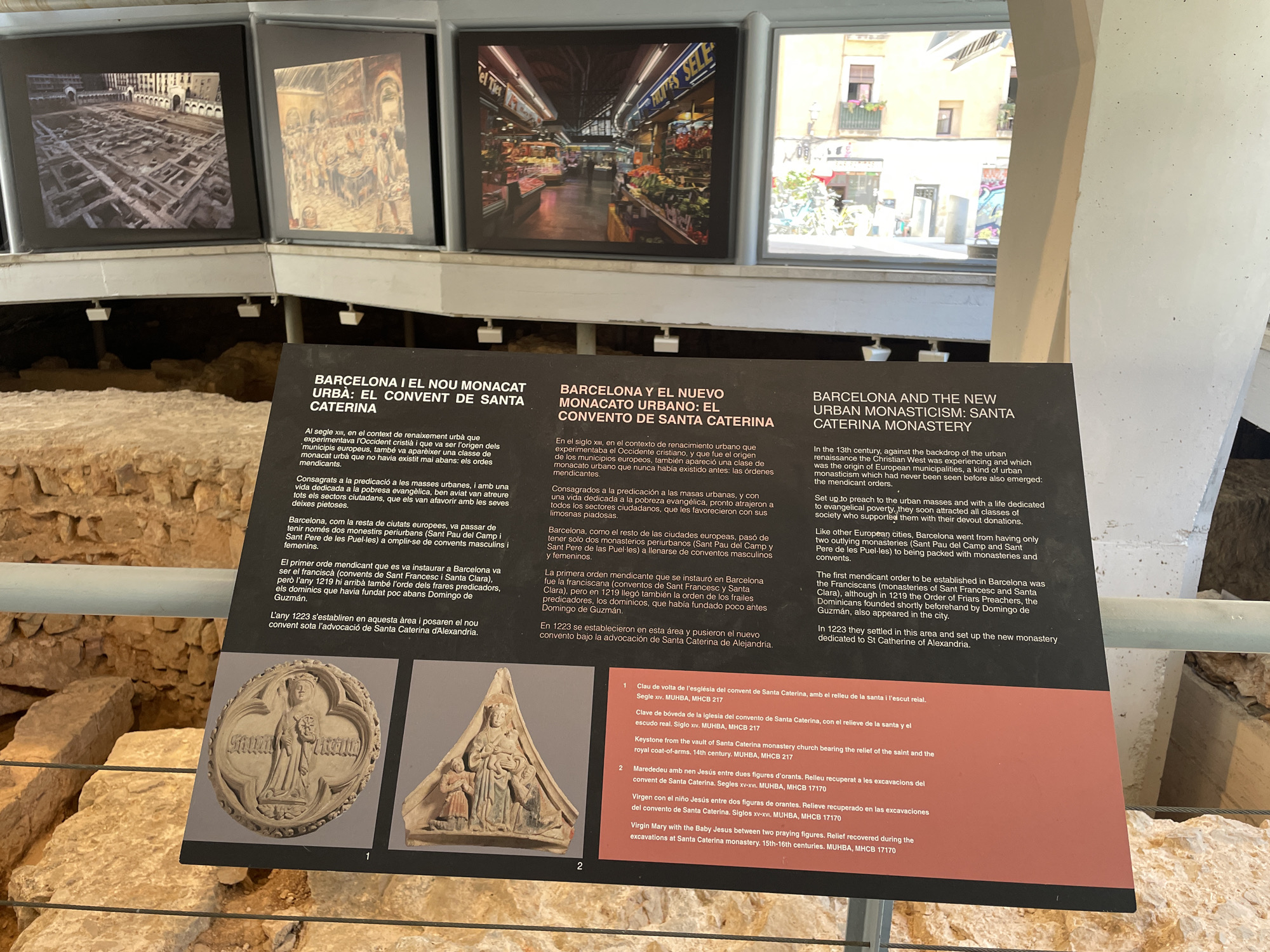 This image captures an indoor museum exhibit focusing on the history of the Santa Caterina Monastery in Barcelona. The primary subject is a dark informational display board featuring text in Catalan, Spanish, and English, detailing the rise of urban monasticism and the establishment of the Santa Caterina monastery in the 13th century.
Above the display board, three framed images or screens provide visual context: the leftmost shows archaeological ruins, possibly the monastery's foundations; the middle depicts a bustling indoor market; and the rightmost is an outdoor street scene with buildings and bicycles.
Below the textual information, two relief sculptures are prominently displayed, with accompanying labels in three languages. The first (labeled '1') is a circular keystone from the monastery's church, dating to the 14th century, depicting a saint and a royal coat-of-arms. The second (labeled '2') is a triangular relief of the Virgin Mary with the Baby Jesus, flanked by two praying figures, recovered from excavations and dated to the 15th-16th centuries. Partially visible beneath the exhibit are actual stone archaeological remains, confirming the setting as an excavation site or a museum built over one.
The visible text discusses the emergence of mendicant orders in 13th-century urban centers, specifically the Franciscans and Dominicans in Barcelona, and the founding of the Santa Caterina monastery in 1223. The museum setting provides historical information and artifacts relating to a significant historical site in Barcelona.