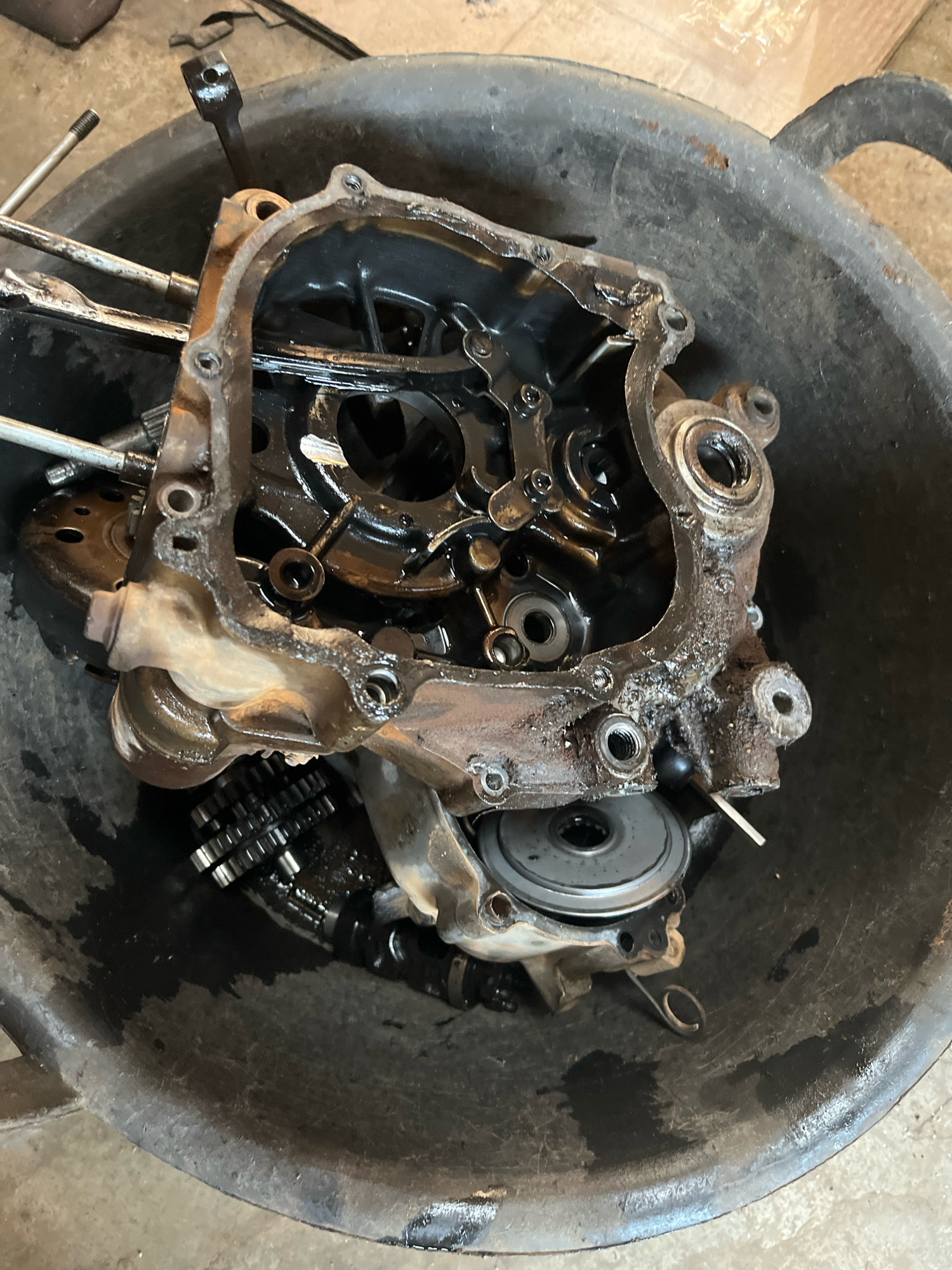 An overhead, close-up shot captures a disassembled motorcycle engine component, likely a gearbox or clutch housing, submerged in a dark, oily basin. The metallic parts are stained with oil and rust, with various gears and internal mechanisms visible. The basin itself is a dark, textured material, showing signs of wear and grime. The overall impression is one of mechanical disrepair or maintenance. The scene is devoid of any contextual clues to suggest its location in Kebumen, Indonesia.