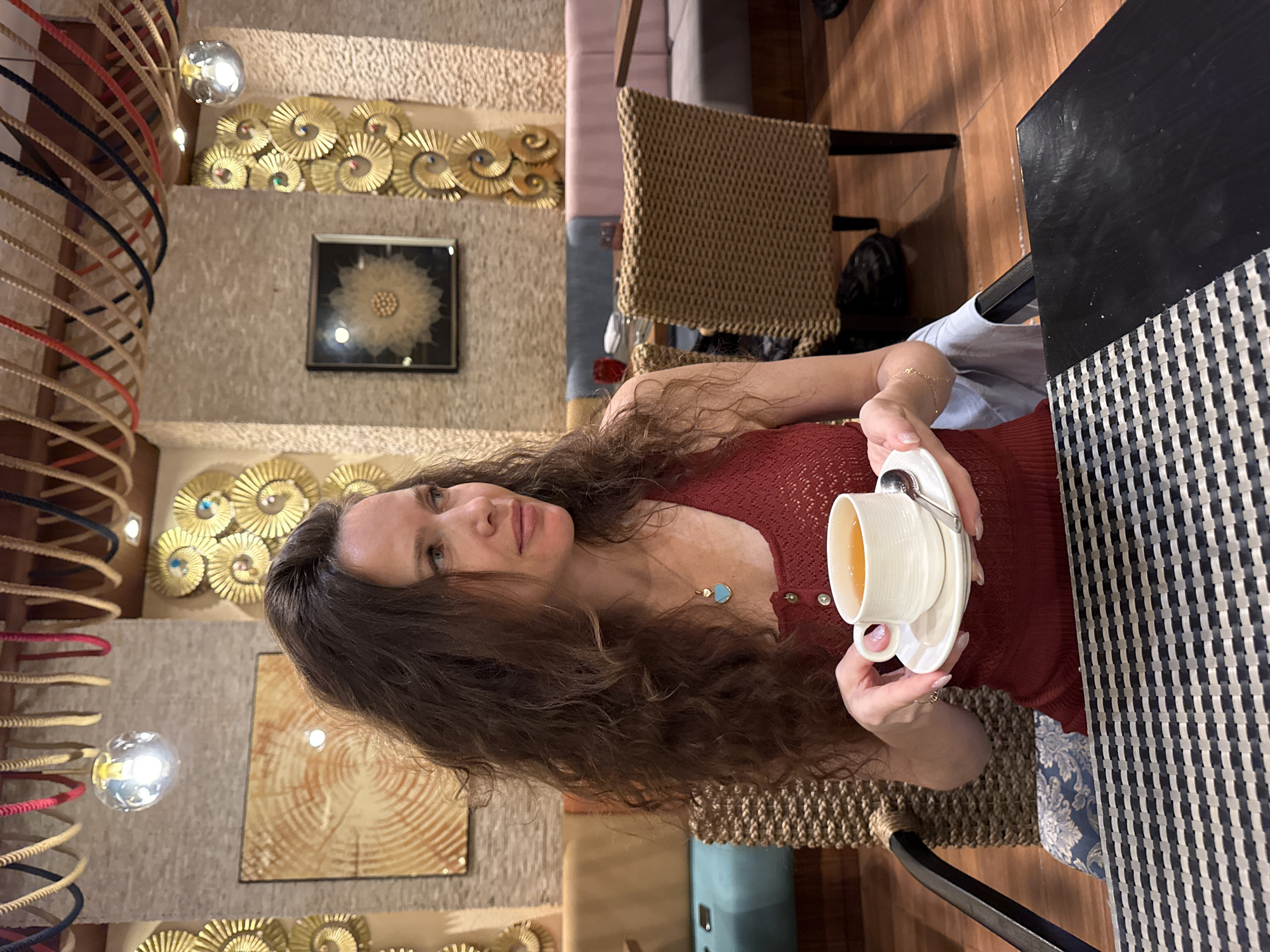 A single adult female is captured in a restaurant or lounge setting, positioned in a three-quarter profile looking toward the right side of the frame with a neutral, slightly pensive expression. She possesses long, dark, wavy hair and wears a reddish-brown, possibly rust-colored, sleeveless top with a knit or crocheted texture and small buttons. A thin gold chain necklace with a light blue, heart-shaped pendant is visible. Her skin displays patches of lighter pigmentation. She holds a white ceramic teacup with an amber liquid and a matching saucer with a small silver spoon; her left hand grips the cup handle, and her right hand supports the saucer.
She is seated at a dark-colored table with a black and light grey/white checkered placemat, in a woven rattan or wicker armchair with a dark metal frame.
The background features an interior with a decorated ceiling traversed by parallel lines of braided ropes in red, beige, dark blue, and black. Glass globe pendant lights and recessed spotlights provide illumination. The textured walls are light-colored and adorned with two framed art pieces: one with concentric circles resembling tree rings, and another displaying an intricate light-colored floral or starburst pattern against a dark background. Gold-colored, circular, spiral-shaped decorative elements, some featuring small colorful centers (blue, green), are vertically mounted on the walls.
Further back, seating includes a light blue upholstered banquette to the left and a light pink upholstered banquette to the right. Another wicker armchair is partially visible facing away from the camera. A red glass rests on a distant table. The flooring is wooden. The lighting is dim and warm.
No visual cues within the image definitively identify the location as Dubai, United Arab Emirates.