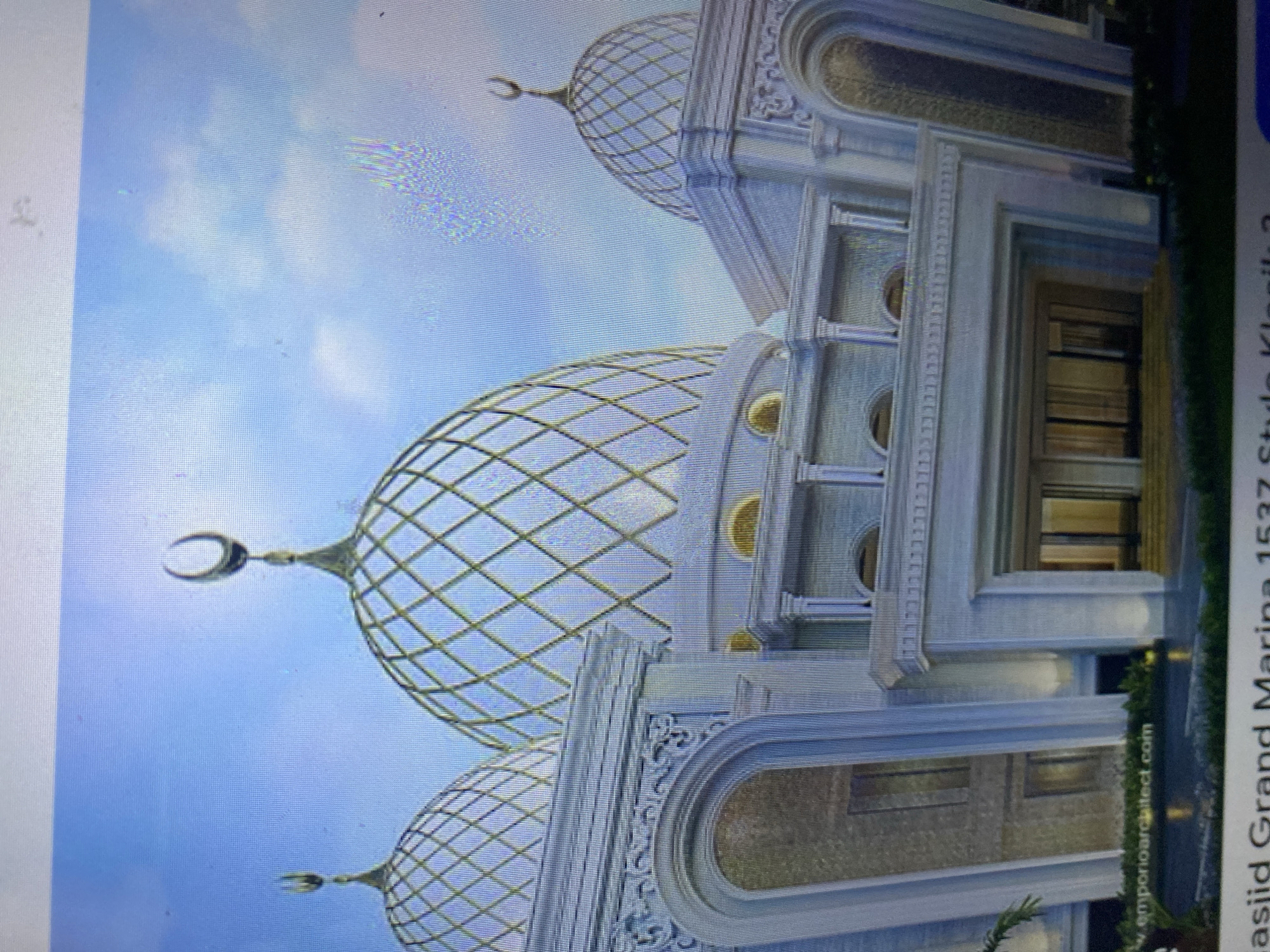 This is a digital rendering of the Grand Marina Mosque located in Ta', Indonesia. The mosque features a modern design with prominent white marble facades and ornate golden domes. The domes are characterized by a lattice pattern and are topped with crescents. The entrance to the mosque is grand, with large archways and detailed stonework. Lush greenery surrounds the base of the building, suggesting a well-maintained landscape. The sky above is a clear blue with subtle cloud formations. The image is watermarked with "www.emporioarchitect.com", indicating the architectural firm responsible for the design. Text at the bottom of the image identifies the project as "Masjid Grand Marina 1537 Style Klasik 2".