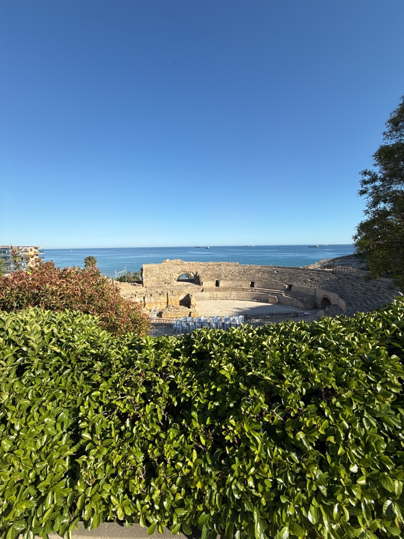 The image shows the Roman Amphitheatre of Tarragona, Spain, overlooking the Mediterranean Sea. The amphitheater's ruins are visible behind a hedge. The sky is clear and blue. Ships are visible on the horizon. Bushes and trees are in the foreground and to the right. A building can be seen in the distance to the left. Seating is setup inside the amphitheater.