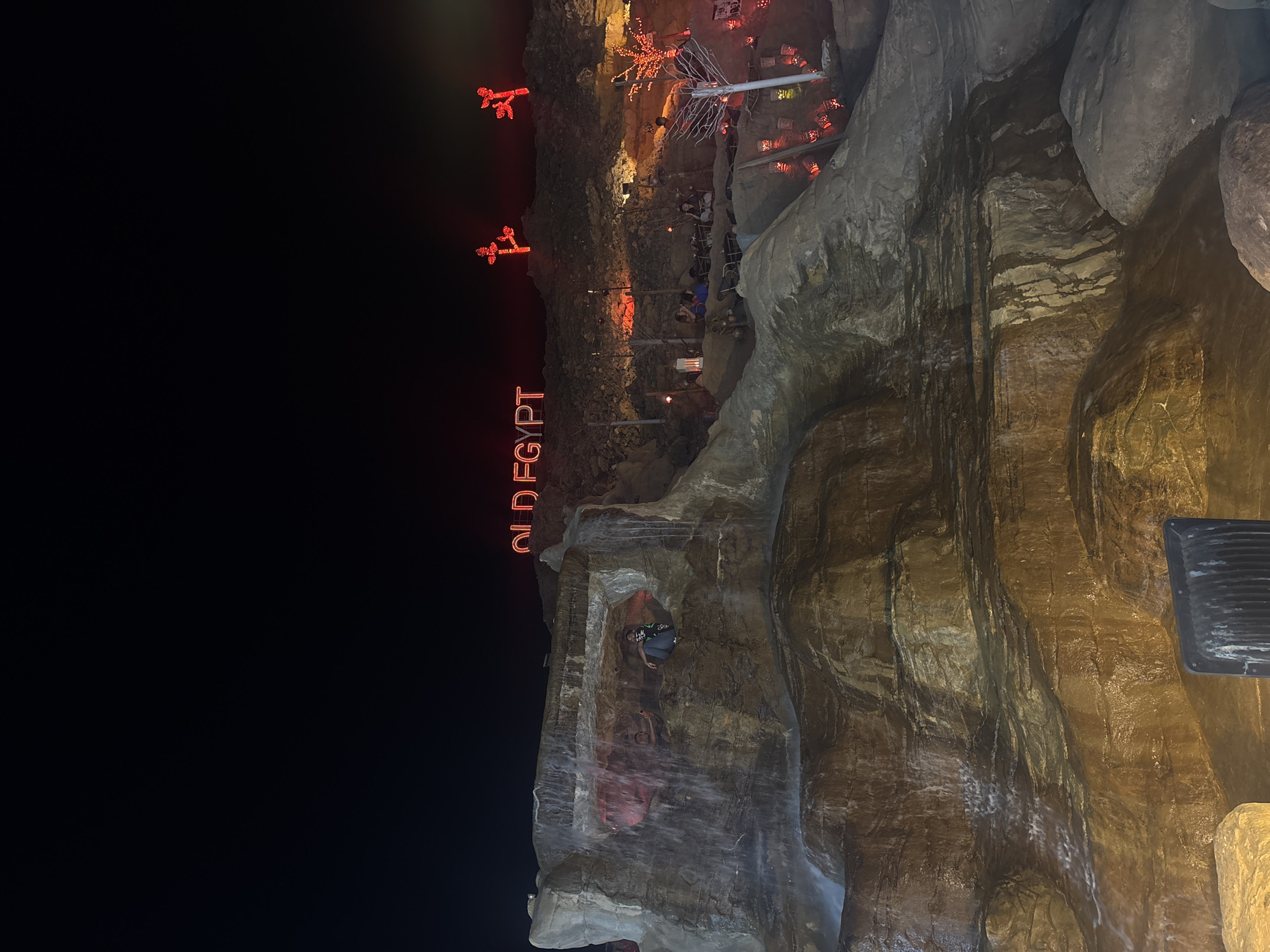 The image depicts a nighttime scene in Sharm El Sheikh, Egypt. In the background, red neon signs spell out "OLD EGYPT". Two additional red, vaguely palm tree-shaped signs are above. A restaurant or bar area is present with tables and people. The foreground features a rock wall with a waterfall. A man is visible inside the rock formation. A black object is mounted on the side of the waterfall.