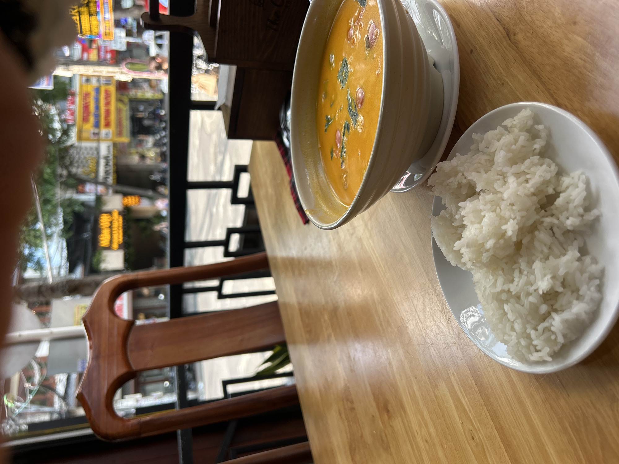 A view from a table in Nha Trang, Vietnam, showing a bowl of tomato soup with herbs and pieces of tomato, served with a side of white rice. The table is wood and the chair is made of dark wood. A window behind the chair shows storefronts in Nha Trang.