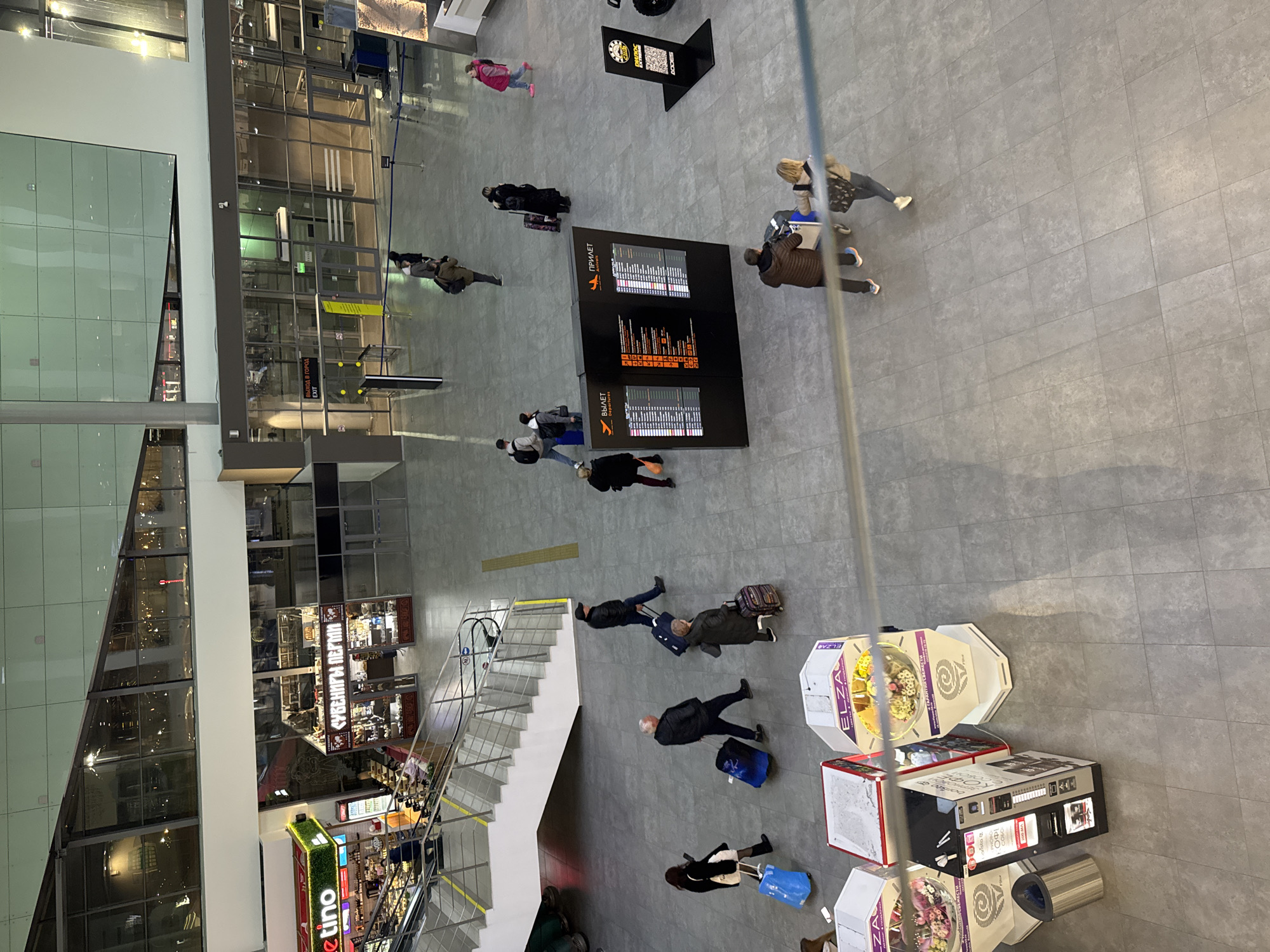 The image depicts an overhead view of a commercial space, likely an airport terminal in Bolshoye Savino, Russia. People, presumably travelers, are seen walking with luggage. Some are gathered near a digital display board showing flight arrivals and departures in Russian. There is a staircase, shops, vending machines and exits to the city. The interior has a modern design with tiled floors.