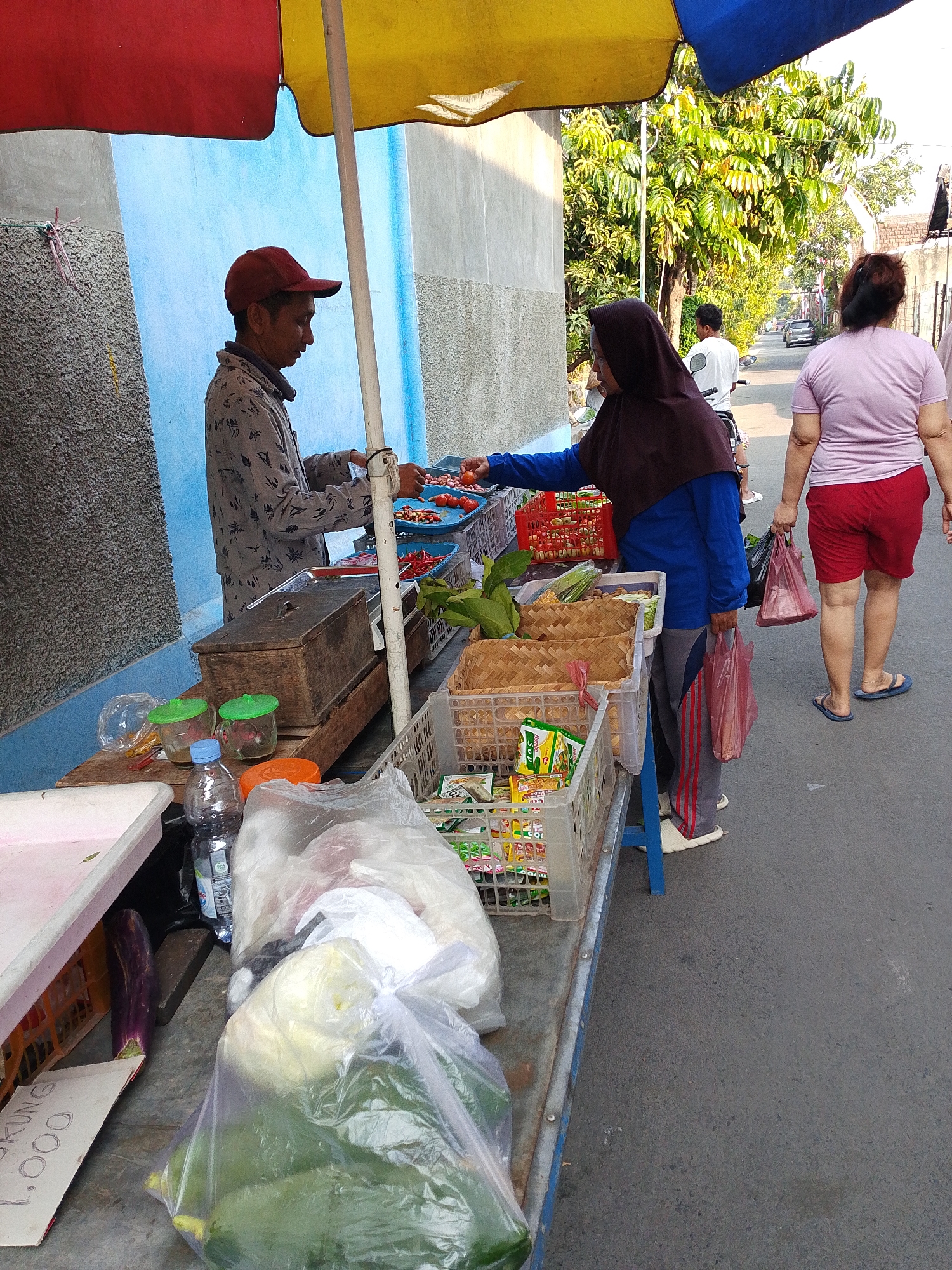 A street vendor sells vegetables from a cart under a blue, red, and yellow awning in Kediri City, Indonesia. The vendor, wearing a red cap and a patterned shirt, serves a customer wearing a dark hijab and a blue jacket. The customer is reaching for a tomato. The cart is filled with various vegetables, including onions, chilies, and leafy greens, along with packaged goods. A woman in a pink shirt and red shorts walks away from the cart, carrying plastic bags. The scene is set on a paved street with trees and buildings in the background. A sign on the cart reads "1.000".