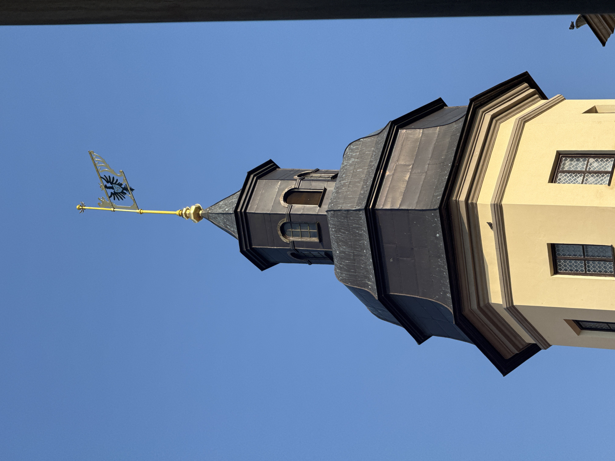The image captures the upper section of a tower, likely part of the Nesvizh Castle complex in Nesvizh, Belarus. The tower has a distinctive architectural style with a light yellow base and a dark, multi-faceted roof. On top of the tower, a golden flagpole extends upwards, supporting a decorative banner with an emblem of a black eagle. The sky is a clear, vibrant blue. The lower portion of the tower shows a couple of windows with decorative grilles. A dark, vertical element is visible on the right side of the frame, partially obscuring the tower.