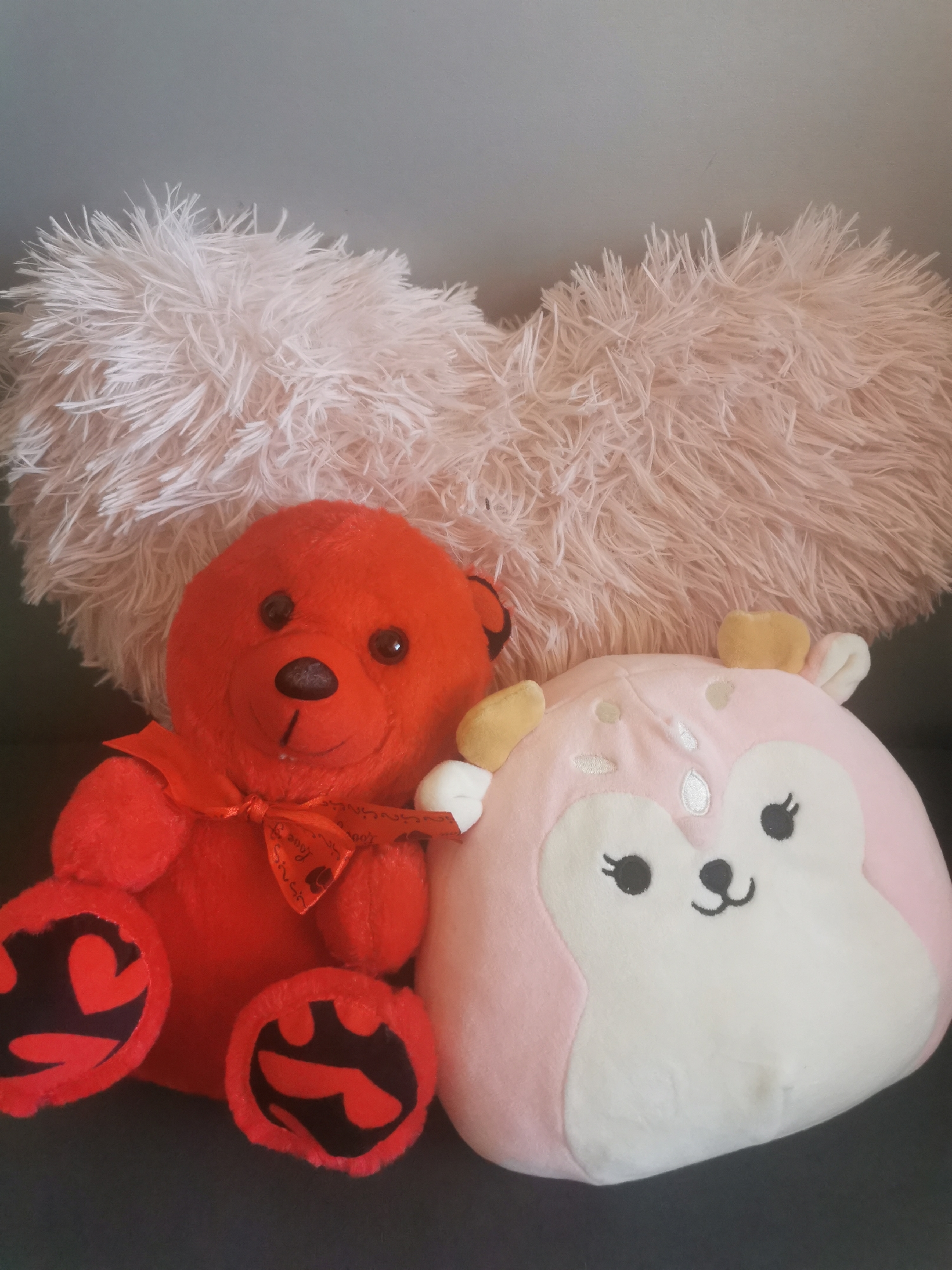 The image captures a cozy arrangement of three plush items positioned indoors against a simple background. In the foreground, two plush toys are seated side-by-side on a dark gray surface, which appears to be upholstery from a couch or chair. Behind them, a large, fluffy, light pink pillow, possibly heart-shaped, serves as a soft backdrop.
On the left, a bright red teddy bear with black button eyes and a black nose is prominently featured. Around its neck is a red ribbon tied in a bow, which bears the repeated white text "Love You So Much." The soles of its feet are decorated with black patches featuring bold red heart shapes.
To the right of the teddy bear sits a round, pink plush toy, consistent with the style of a Squishmallow, resembling a fawn or similar animal. It has a white face with embroidered black eyes, eyelashes, and a small, friendly black nose and mouth. Small yellow antlers and white ears extend from its pink head.
The background above the fluffy pillow reveals a plain light gray wall. The lighting in the scene is soft and even, characteristic of an indoor setting, likely during daylight hours. No specific activity is taking place other than the static display of the plush toys. The overall scene conveys a feeling of comfort and cuteness. No specific location cues for Blagoevgrad, Bulgaria, are discernible from the image content itself.
