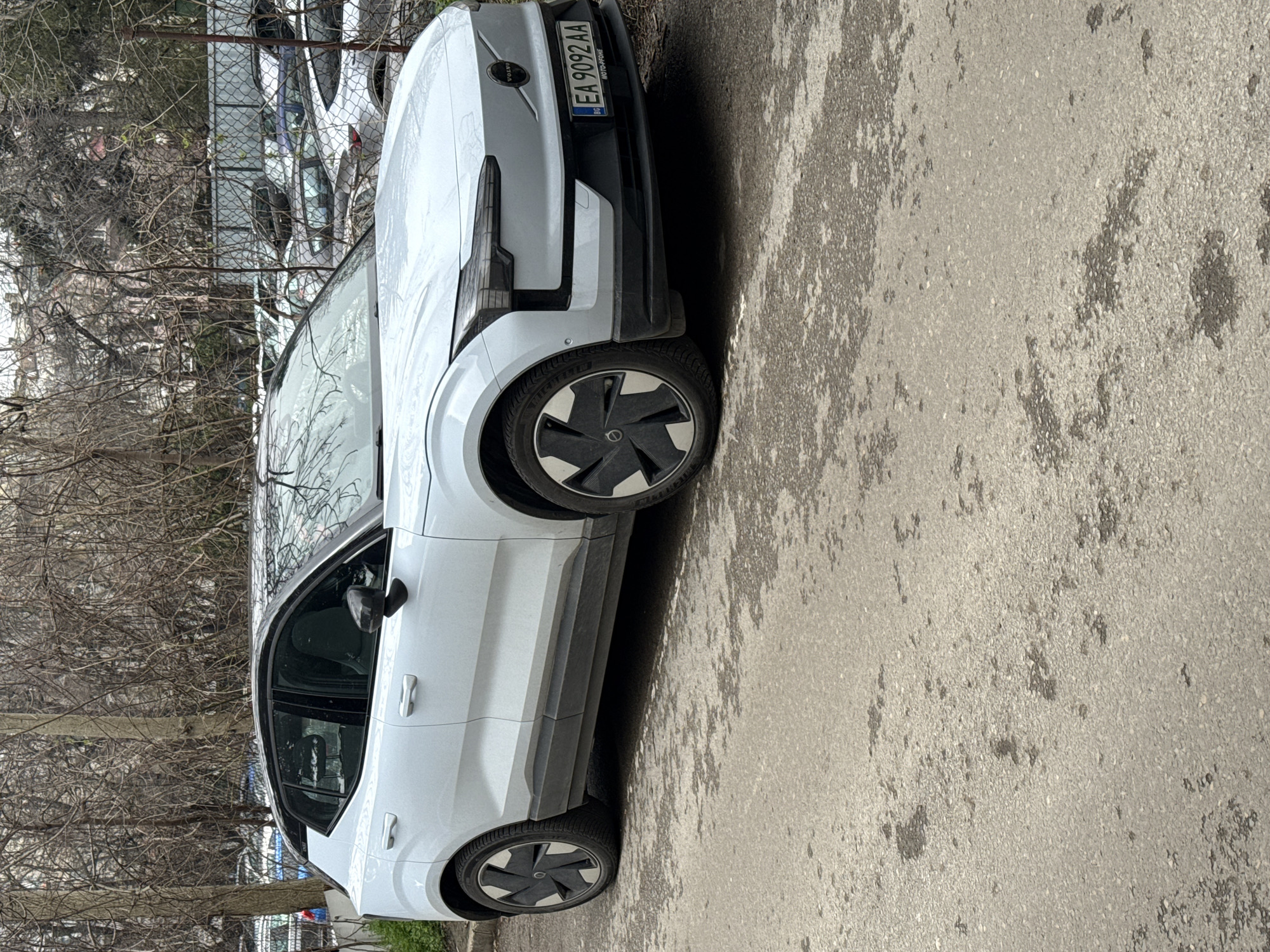 A white Volvo XC40 Recharge is parked on an asphalt surface next to a sidewalk. The car's Bulgarian license plate reads "EA 9092 AA". Behind the car is a metal fence with other parked cars visible behind it. Bare trees are present in the background, suggesting it might be winter or early spring. The overall scene appears to be a typical urban street setting, possibly in a residential or commercial area. Though the exact location cannot be determined, the number plate indicates that the car is in Varna, Bulgaria.