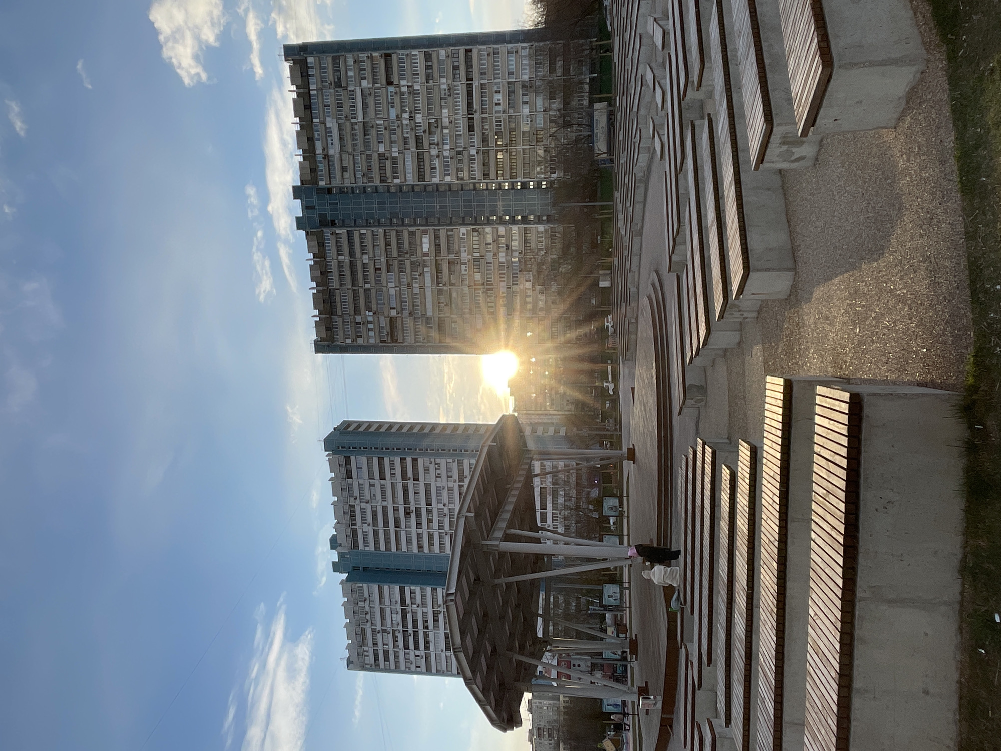 The image depicts a city scene in Moscow, Russia. Two tall residential buildings with multiple floors and numerous windows stand in the background. The sun is positioned between the buildings, creating a bright glare.
In the foreground, there's a public space with benches made of wood and concrete, arranged in a parallel fashion. A covered structure or pavilion is present.
Two people are visible in the middle ground of the image. The sky is blue with scattered clouds.