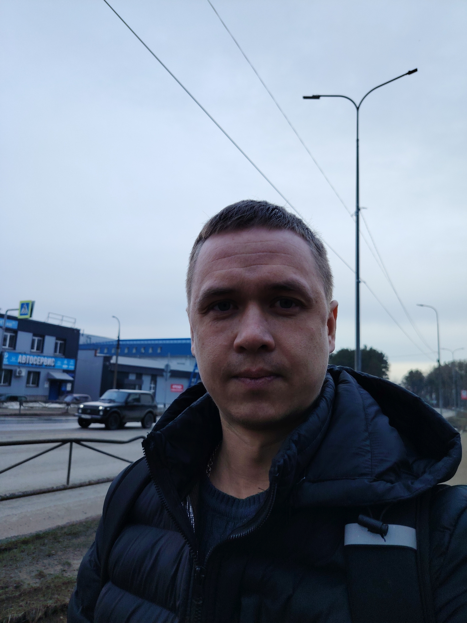 A man is taking a selfie outdoors in Chaikovsky, Russia. He is wearing a black puffer jacket and a gray sweater. In the background, there is a two-story building with "АВТОСЕРВИС" (Auto Service) written on it in Russian. A black SUV is driving on the road in front of the building. Overhead power lines and streetlights are visible against a cloudy sky. A concrete barrier is in the foreground, suggesting he is standing on a sidewalk or pedestrian area.