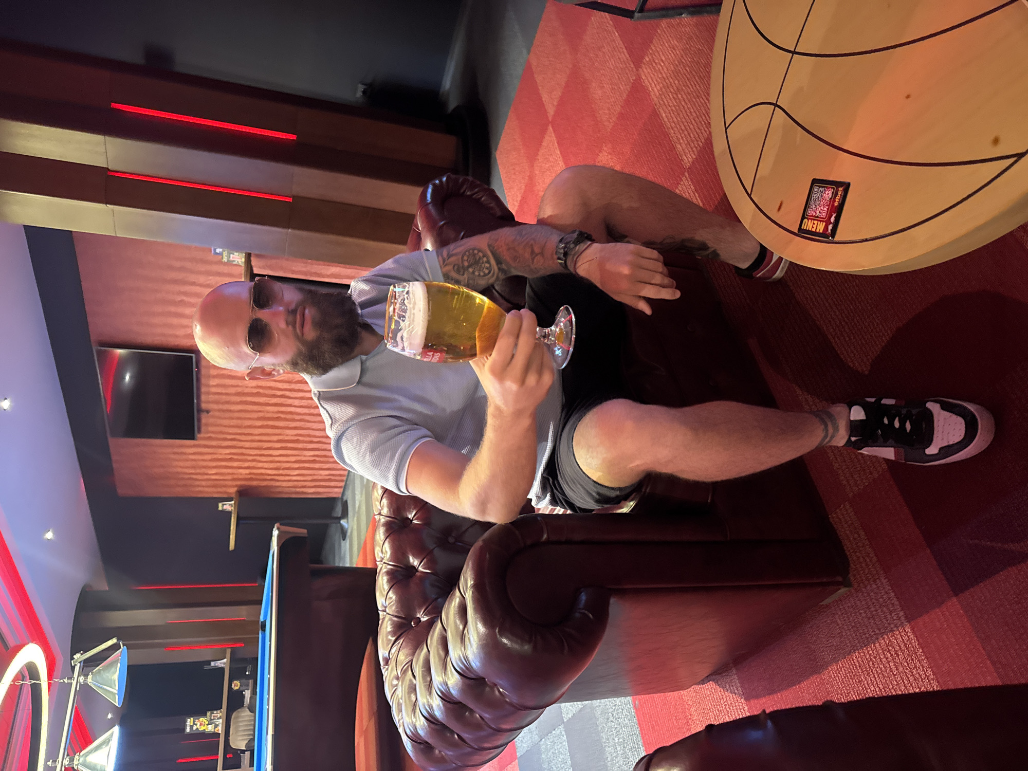 A bald man with a beard is sitting in a leather armchair, holding a glass of beer. He is wearing sunglasses, a light blue collared shirt, and dark shorts. His arms are tattooed. He is wearing black and white sneakers with red striped socks. He is seated in a room with a red patterned carpet. There is a round wooden table with a menu beside him. A pool table and bar area are visible in the background. Red lighting fixtures illuminate the space. The location is a bar or lounge in Sofia, Bulgaria.