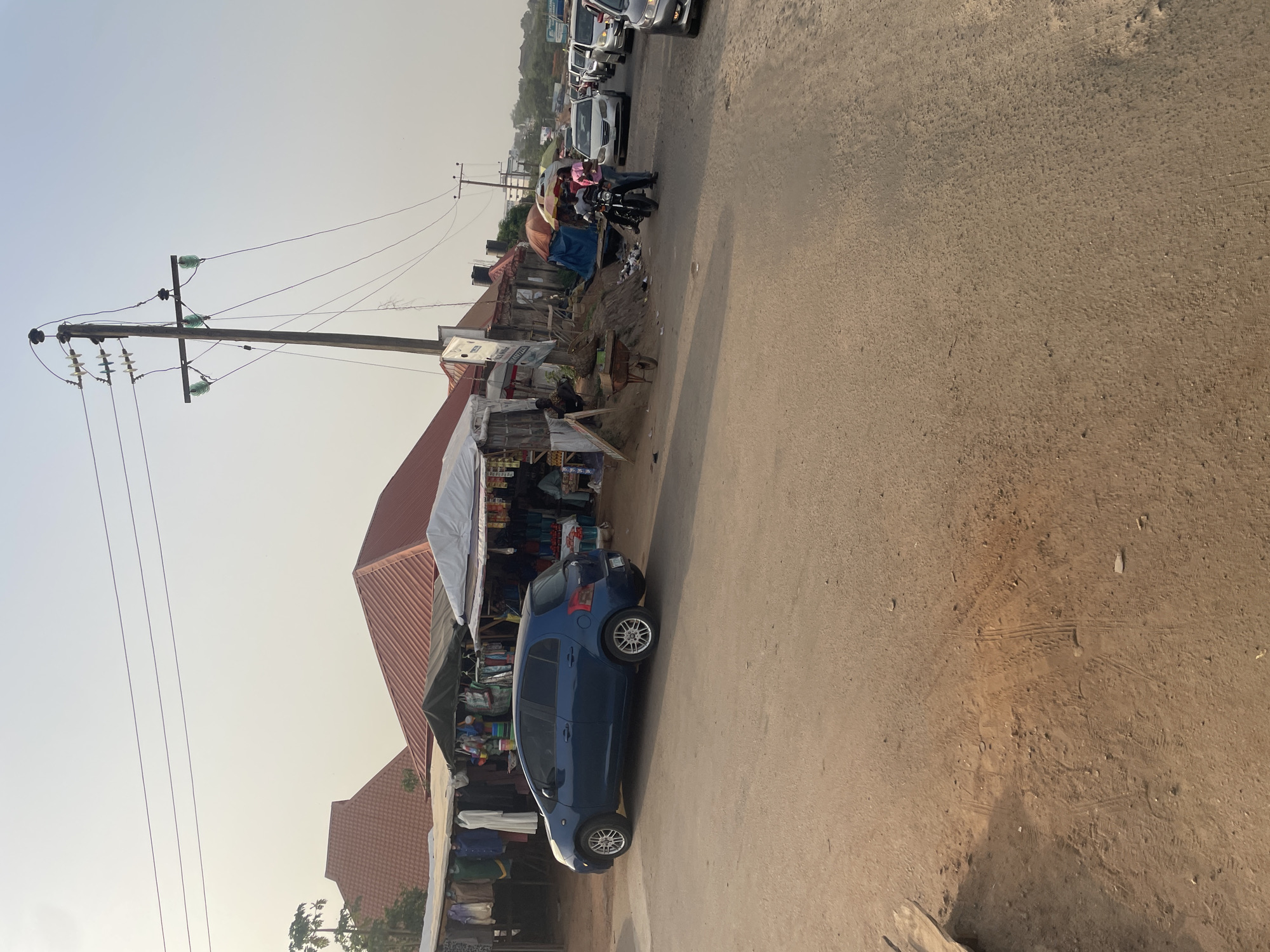 The image captures a bustling street scene, likely a market area, in Abuja, Nigeria. The foreground is dominated by an unpaved, sandy-brown road, showing tire marks and scattered debris. On the left side of this road, a blue hatchback car, possibly a Toyota Yaris, is parked beside a row of market stalls.
These stalls are rudimentary, featuring structures with white and dark tarpaulin canopies, under which various goods are displayed. Several people are visible around these stalls, engaged in daily activities, suggesting vending or shopping. A tall wooden utility pole, equipped with multiple power lines and green glass insulators, stands prominently behind the market stalls.
Further down the road, in the mid-ground, a person on a motorcycle, potentially with a passenger, is seen under an open umbrella, presumably for sun protection. A line of other vehicles, including white and silver cars, are parked along the road. In the background, more buildings with reddish-brown roofs are visible, along with distant structures or hills under a bright, clear daytime sky. The lighting is soft and even, suggesting a morning or late afternoon setting. On a white signboard near one of the market stalls, the word "AVAILABLE" is discernible. The overall scene depicts a lively local commercial and transport hub.
