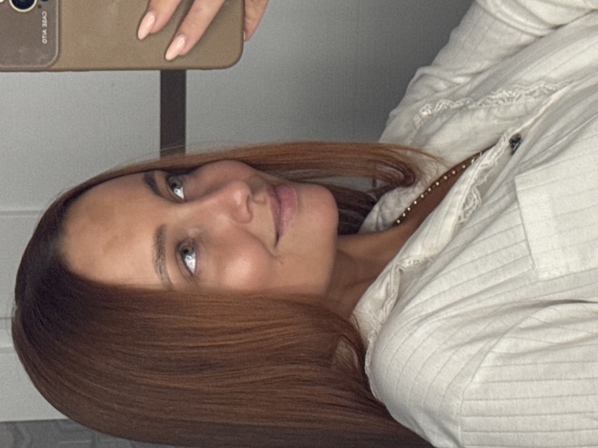 A medium shot captures a woman with reddish-brown hair styled straight, looking to her right and slightly upwards, in what appears to be a mirror selfie. She is wearing a light-colored, collared shirt with subtle vertical pinstripes and a visible button. A thin, gold chain necklace is also apparent. Her right hand is visible, holding a beige phone case. The background is a plain, light gray wall. No specific landmarks or indications of Chaikovsky, Russia are present in the image.