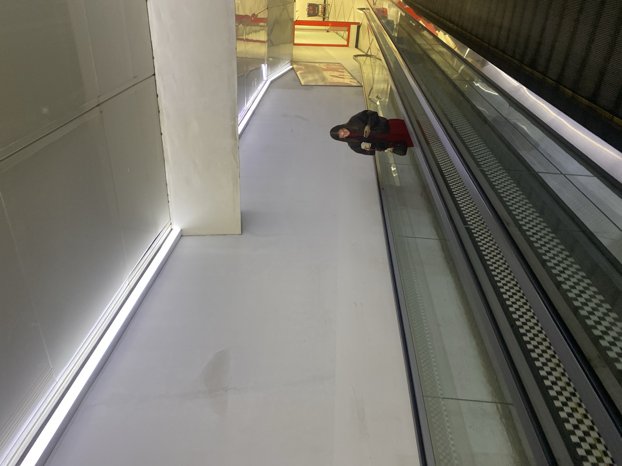 A woman is standing on a moving walkway in a modern, brightly lit interior space. She is wearing a dark jacket, a red dress, and has a cup in her hand. The moving walkway is next to an escalator. The wall is white, with a bright linear light fixture. A red and white geometric shape is visible in the background. The location is Yekaterinburg, Russia.