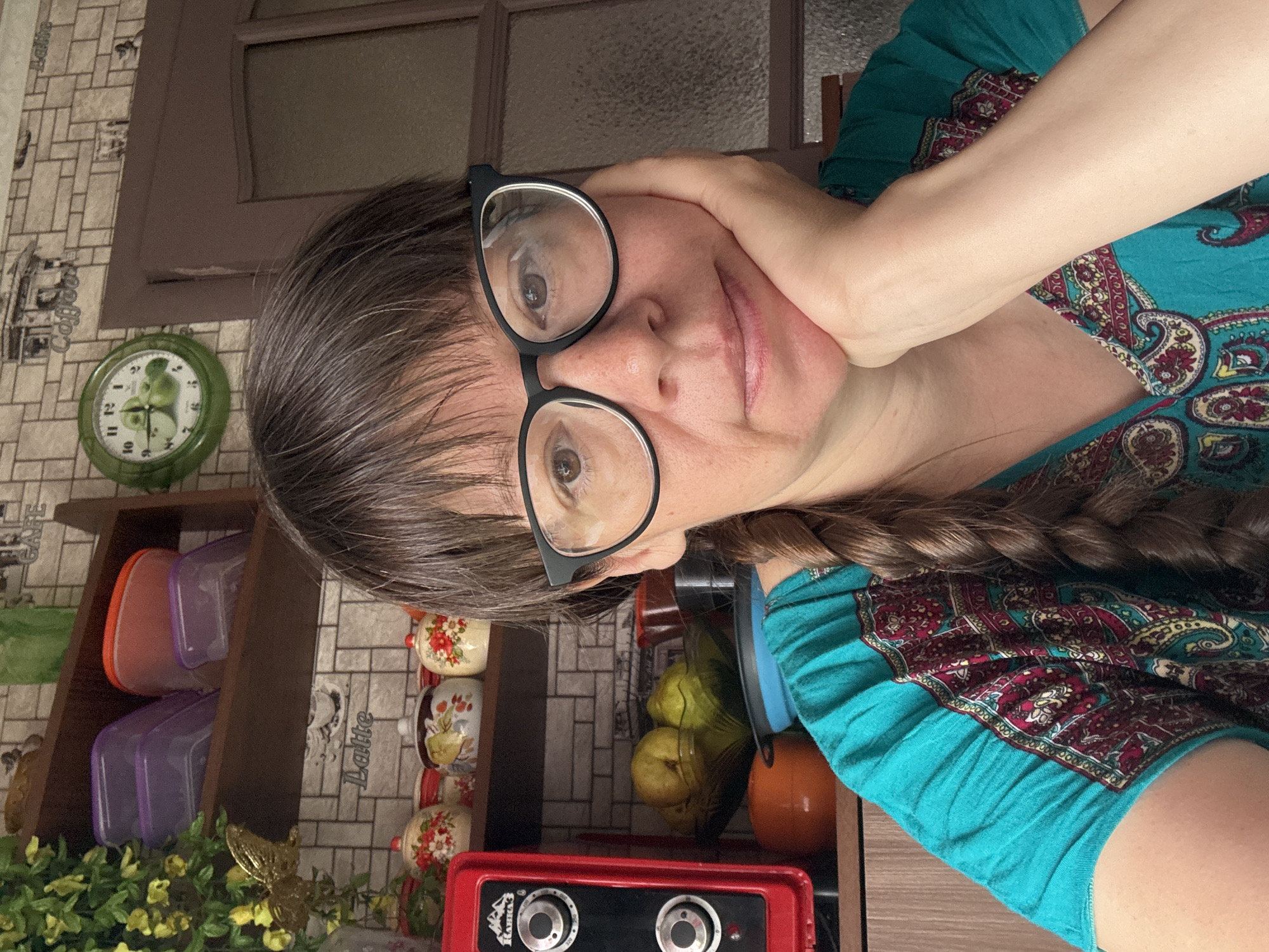 A middle-aged Caucasian woman with brown hair and glasses is shown from the chest up, resting her chin on her right hand. She is wearing a teal top with a paisley pattern. The background includes a kitchen shelf with various containers and jars, a red appliance, a bowl of fruit, and a wall clock. The setting appears to be a domestic environment, likely a kitchen, within the city of Vetluzhsky, Russia.