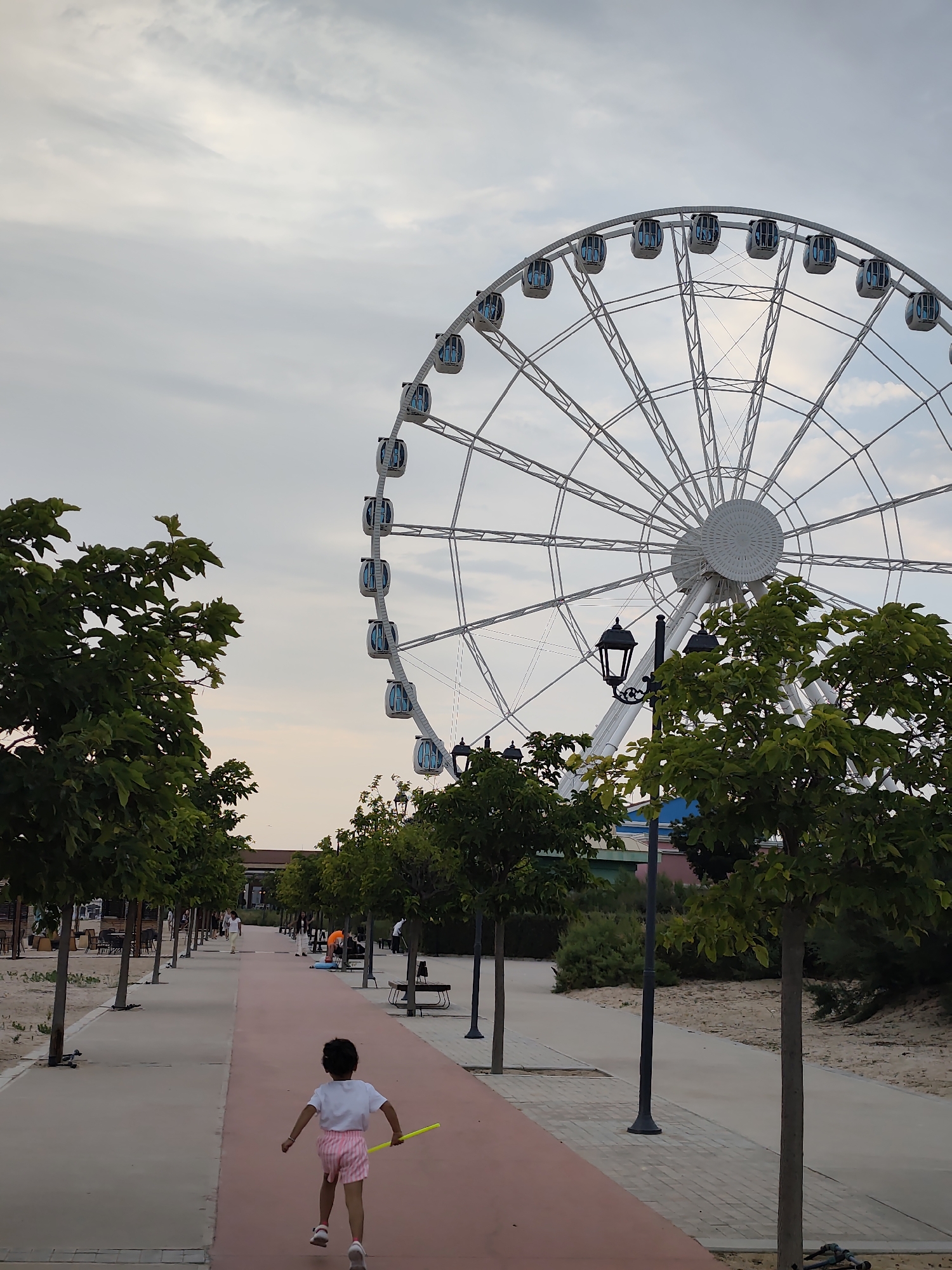 A young child wearing a white t-shirt and pink striped shorts runs down a red pathway in an outdoor public space, holding a yellow object. To the right of the path, trees are planted in neat rows. In the background, a large white Ferris wheel with blue-tinted cabins dominates the scene under a cloudy sky. The area appears to be a promenade or park, with benches and lampposts visible. The location is identified as the Aktau Ferris Wheel area in Aktau, Kazakhstan.