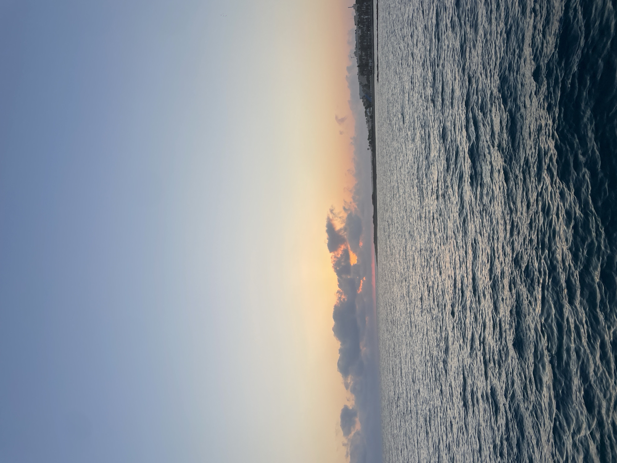 The image depicts a serene seascape with a coastal town in the distance. The sky is a gradient of soft blues and oranges, suggesting either sunrise or sunset. A line of clouds stretches across the horizon, partially obscuring the sun which casts a warm glow. The sea is a deep blue with gentle ripples. On the right side of the frame, a coastline is visible, characterized by buildings and vegetation, consistent with the appearance of Yeroskipou, Cyprus. The town appears to be nestled along the shore, with structures that suggest a developed area. There are no people or visible actions in the scene. The overall impression is one of natural beauty and tranquility.