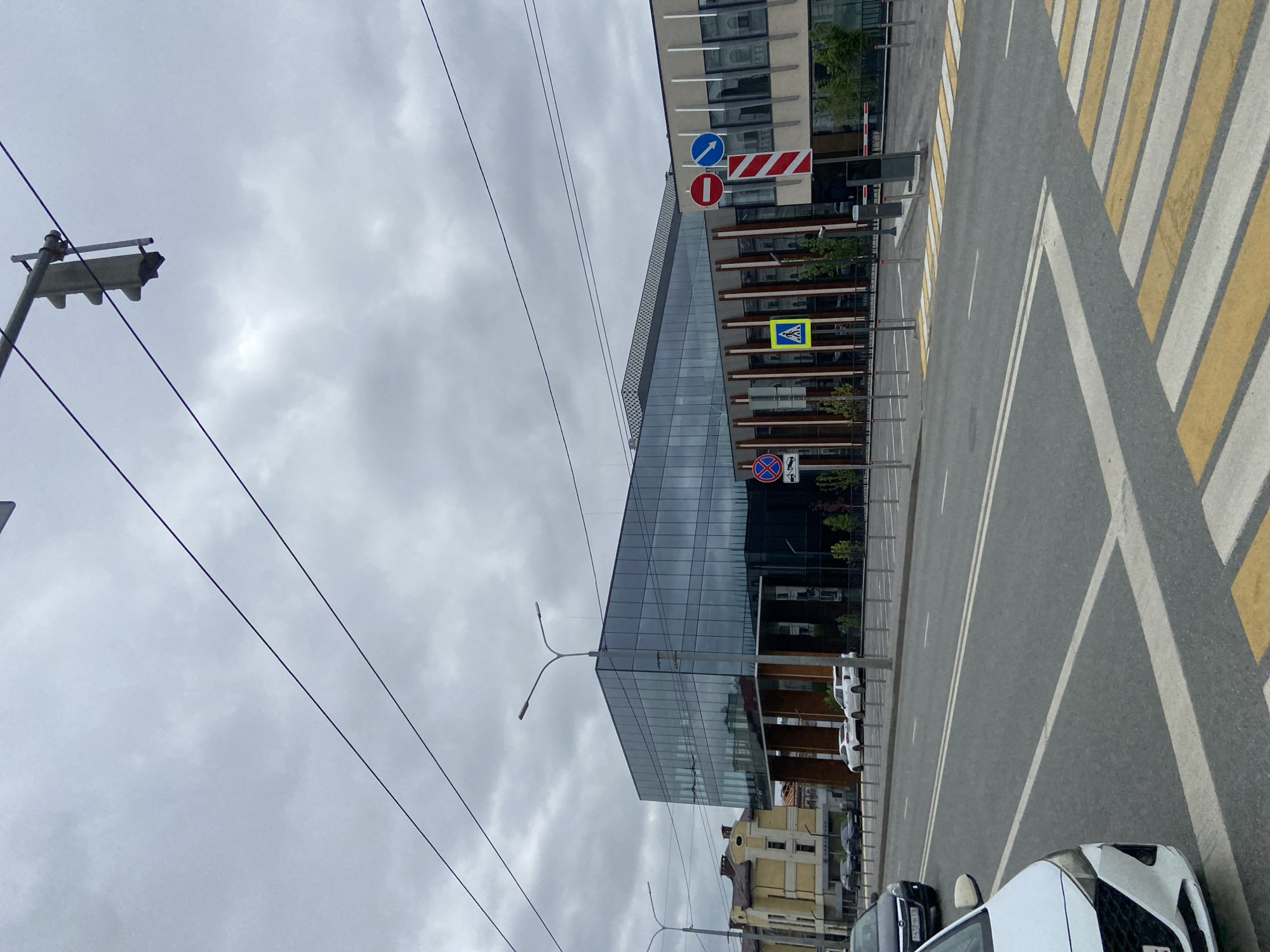This image captures a cloudy daytime street scene in an urban environment, likely Kazan, Russia, as indicated by the context.
The scene is dominated by a large, modern building on the left, featuring a sleek, reflective glass facade that mirrors the cloudy sky and surrounding structures. Behind it, a portion of an older, lighter-colored building with traditional architecture is visible.
To the right and in the foreground, an asphalt road with distinct white lane markings stretches across the frame. A prominent pedestrian crossing, characterized by alternating yellow and white stripes, is visible on the road surface. Several vehicles are present: the front of a white car occupies the immediate foreground on the right, and other cars are parked or moving along the road next to the modern building.
Various road signs are clearly visible, indicating traffic regulations. These include a blue circular sign with a white arrow (mandatory straight ahead), a red circular sign with a white horizontal bar (no entry), and a rectangular sign with red and white diagonal stripes (indicating a barrier or road works ahead). Further down, a yellow square sign with a black pedestrian symbol (pedestrian crossing ahead) is mounted, along with a blue circular sign with a red diagonal line over a car (no stopping), which has a small, unreadable supplementary panel below it.
Overhead, numerous power lines crisscross the overcast sky, and a traffic light pole is partially visible in the upper left corner. The weather is uniformly cloudy, suggesting a diffused light rather than direct sunlight. No human activity is overtly depicted, and no specific text is clearly readable on any signs or vehicle license plates.