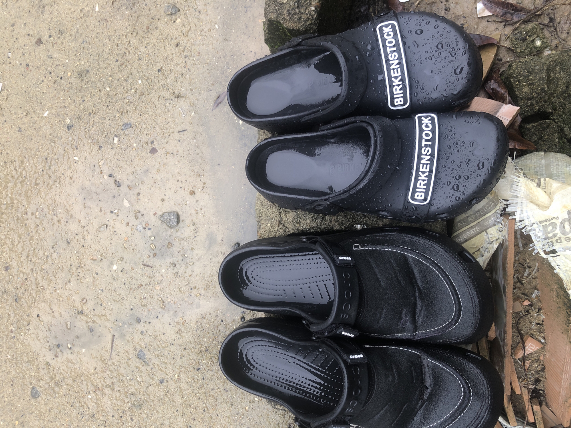 The image shows two pairs of black clogs, one brand is Birkenstock and the other brand is Crocs. Both pairs of shoes appear wet, with water droplets visible on the upper surfaces. The shoes are positioned on an outdoor, wet surface of concrete and debris (possibly tiles and garbage), implying a recent rain. Ado, Nigeria is not directly apparent.