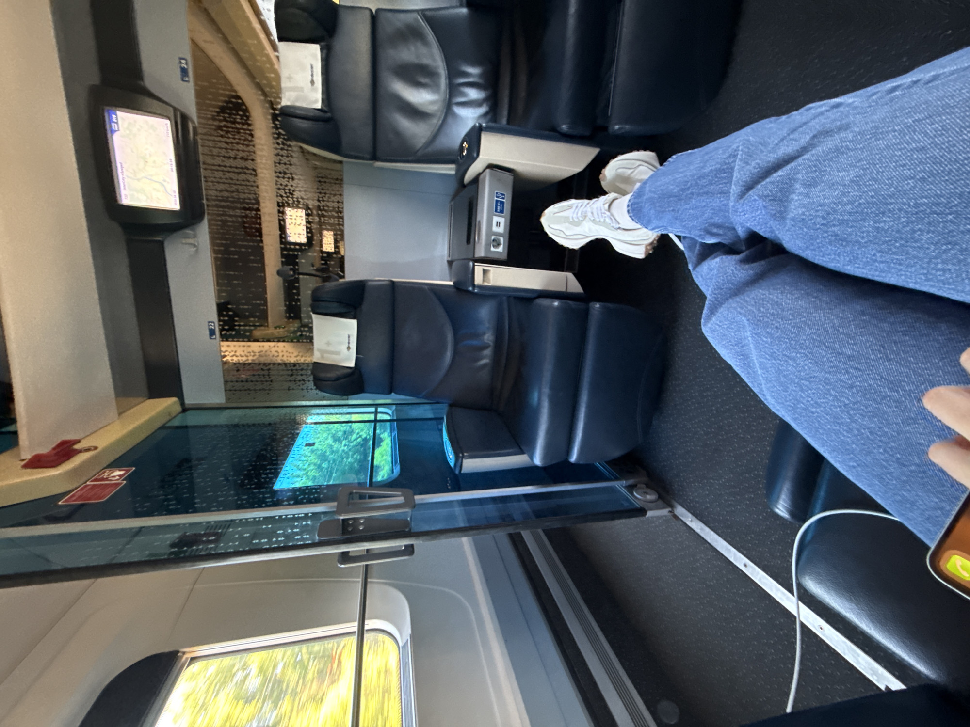 The image captures the interior of a modern passenger train carriage, viewed from a passenger's perspective. In the foreground, a person's legs are visible, clad in blue jeans and white sneakers, crossed at the ankles. Their right hand is partially seen holding a smartphone, connected by a white charging cable.
The train features dark blue, upholstered seats, some with small white headrest covers bearing a subtle logo. Between two seats, a console provides power outlets, including a European-style Type F socket and USB ports. On a grey pillar opposite the seats, a sleek, vertically oriented screen displays a map, likely tracking the train's journey, with "7:30" and "215 km" visible. Text on the screen also partially shows "Departure". Below this screen, the number "L22" is visible, with "L7" also appearing higher up the pillar, likely indicating seat or row numbers.
A large glass divider or partition runs along the side of the carriage, separating the seating area. This divider has a textured, reflective upper section and a clear lower section. Below the divider, a window offers a view of vibrant green foliage outside, indicating the train is in motion through a natural landscape. The scene appears to be set during the daytime, given the brightness outside. The floor is dark gray with a subtle textured pattern. The overall impression is one of comfortable and well-equipped travel.