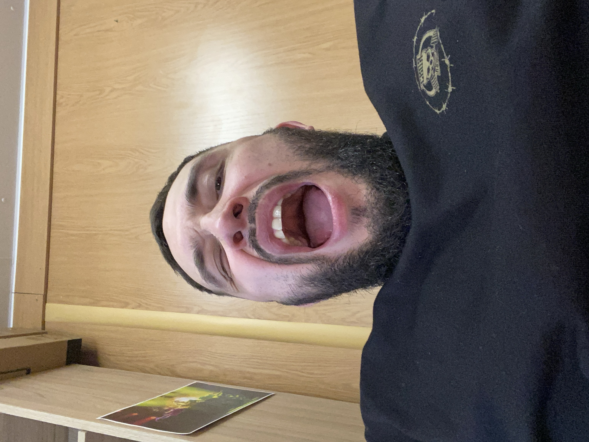 A person with a beard and black hoodie, standing in front of a wooden door and shelf. The person has their mouth open wide. A picture is on the shelf. The image contains no information about the city of Luzhitsy, Russia.