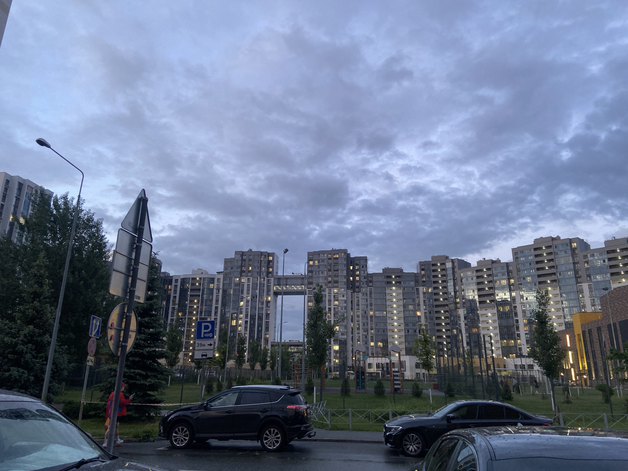 The image captures a modern residential street scene at dusk or dawn, under an overcast sky filled with dense, gray clouds. The wet ground indicates recent rainfall.
In the background, numerous tall, multi-story apartment buildings dominate the skyline. These contemporary structures feature many windows, with artificial lights illuminating various rooms, contrasting with the dim natural light. One prominent building in the center has a large, rectangular architectural cutout extending through its lower section.
The mid-ground features a paved street or road, where several vehicles are visible. Parts of a car are in the immediate foreground, a dark SUV is parked on the left, and another dark sedan is parked on the right. A low metal fence separates the street from a grassy area dotted with young trees and bushes. Faintly visible behind the fence are structures resembling a playground or recreational equipment.
On the left, a streetlamp with a curved arm stands tall, alongside a pole bearing multiple road signs. A clear blue rectangular sign displays a white 'P' for parking, with the text "39m ->" and a car icon below it, indicating parking 39 meters ahead. Another blue square sign with a white right-pointing arrow is also visible.
A single person is observed walking in the mid-left area, wearing a red jacket or top and appearing to be engrossed with a mobile phone. The scene conveys a quiet, typical evening or early morning atmosphere in a newly developed or modern urban residential complex.