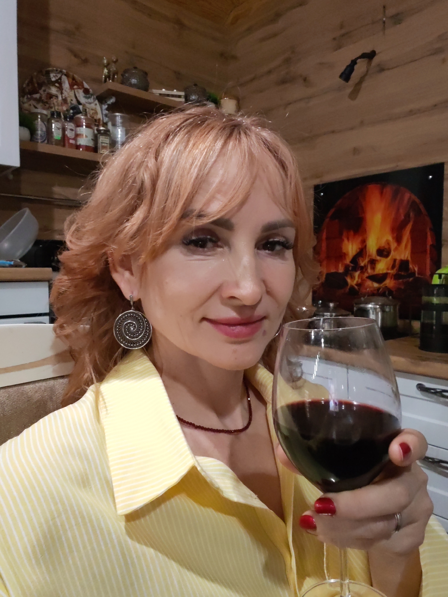 A woman holds a glass of red wine in a kitchen setting in Старое Михайловское, Russia. She has fair skin, red-blonde curly hair with bangs, and wears a yellow and white striped collared shirt, round silver earrings, and a burgundy beaded necklace. Her fingernails are painted red. Behind her, a shelf holds various jars and containers, while a picture of a burning fire hangs on the wall. The walls and ceiling appear to be paneled with wood.
