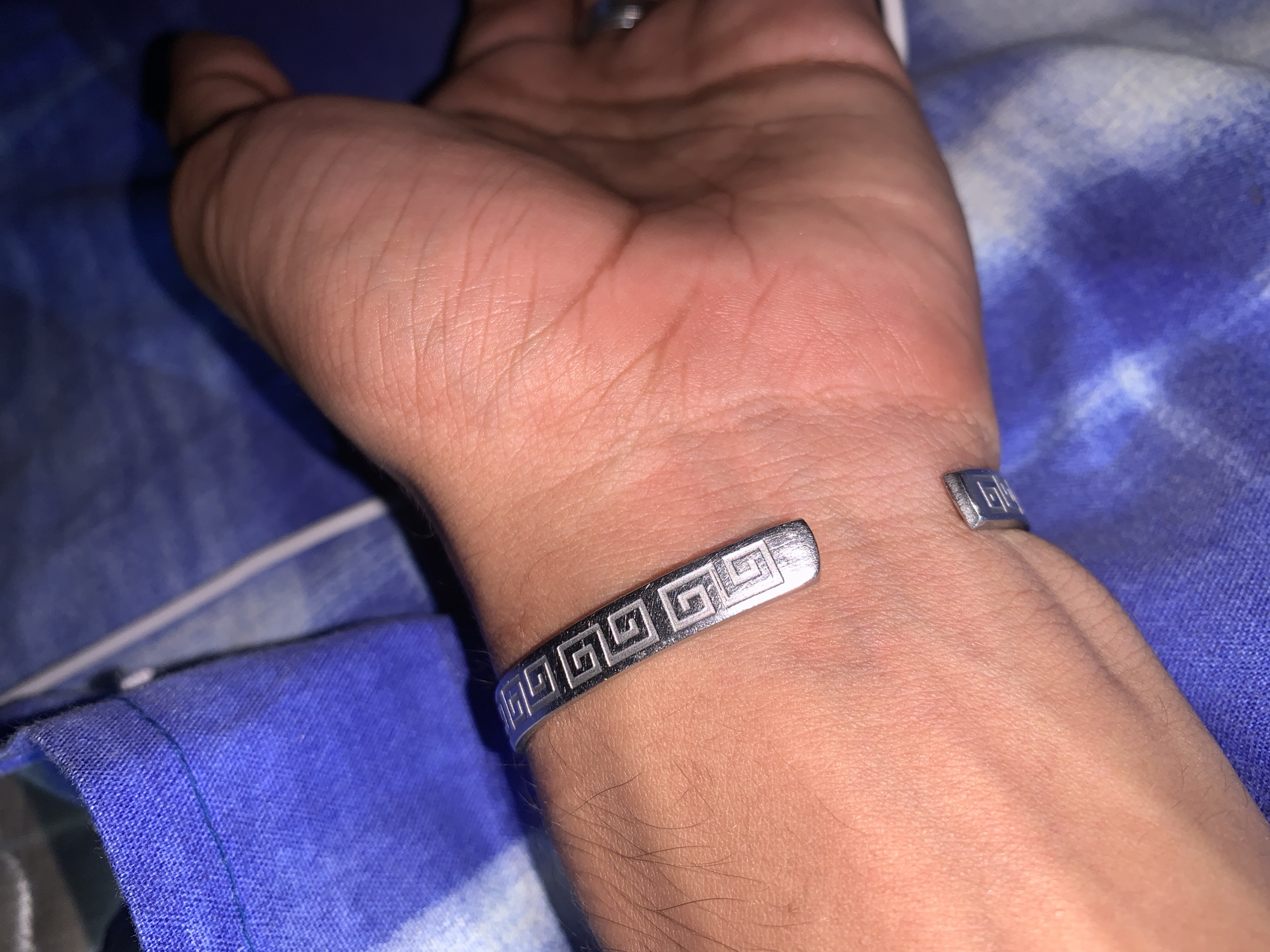 An image captured in Santa Ana, Venezuela, features a human hand, likely an adult's, resting on a blue and white tie-dye patterned fabric, possibly bedding or clothing. The hand's skin tone is medium to dark, with visible lines on the palm and some hair on the wrist and forearm.
A prominent silver-toned cuff bracelet is worn around the wrist. The bracelet features an engraved or embossed geometric design, specifically a repeating Greek key or meander pattern composed of interconnected square spirals. The bracelet appears to be open or has a gap on one side.
In the background, a finger with a faint ring is visible, and the overall lighting suggests an indoor setting, possibly during the evening or in low light, creating soft shadows. No specific activity is depicted; the hand appears to be at rest. There is no visible text in the image.
