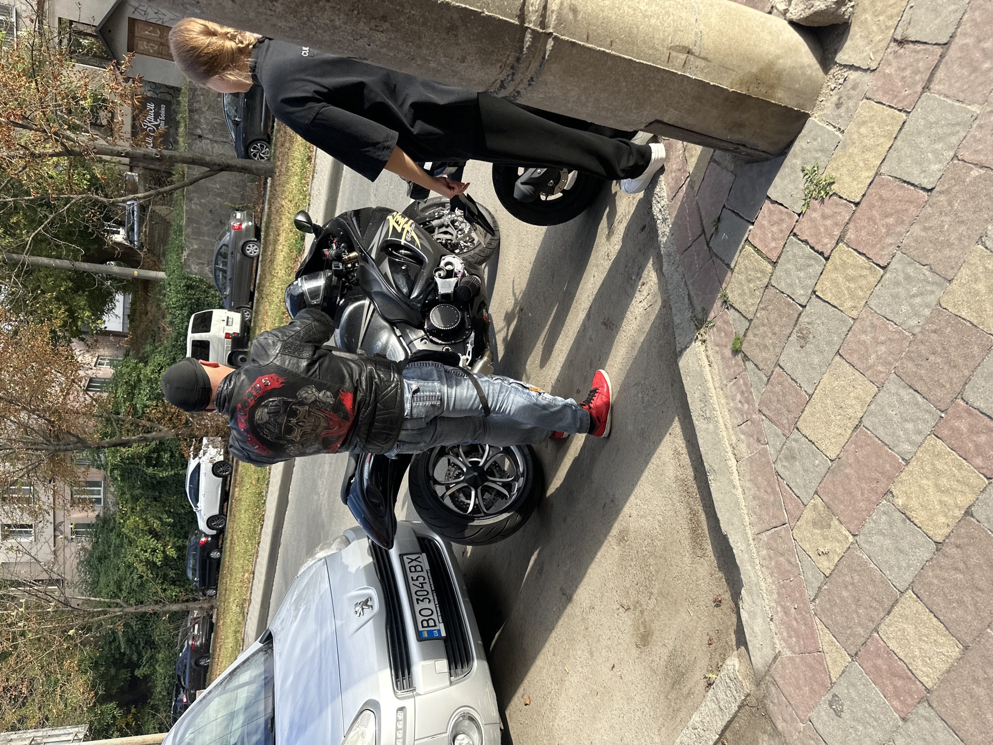A man wearing a black leather jacket with a skull design on the back, a black skullcap, blue jeans, and red sneakers stands next to a black motorcycle. He is looking at the motorcycle. To his left is a silver Peugeot car with the license plate BO 3045 BX. To the man's right, a woman wearing a black t-shirt and black pants stands with her back to the camera, also looking at the motorcycle. The scene takes place on a street in Ternopil, Ukraine, with buildings and trees visible in the background. The ground is paved with cobblestones.