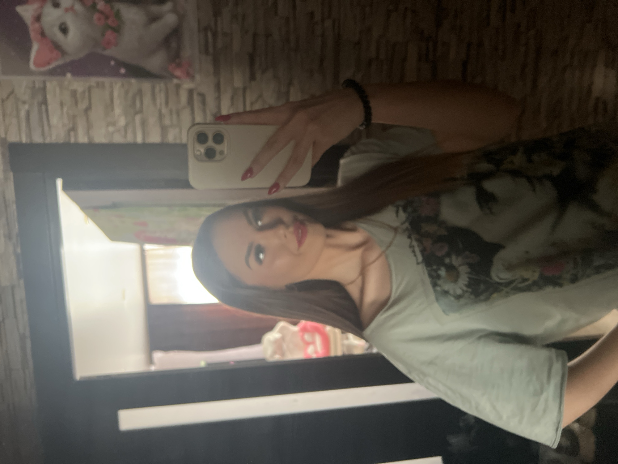 A woman is taking a selfie in a room that appears to be a bathroom. She is holding a smartphone with multiple lenses. The background includes a mirrored surface, a cat figurine, and a tiled wall. The location in the city of Noviy, Russia, is unknown.