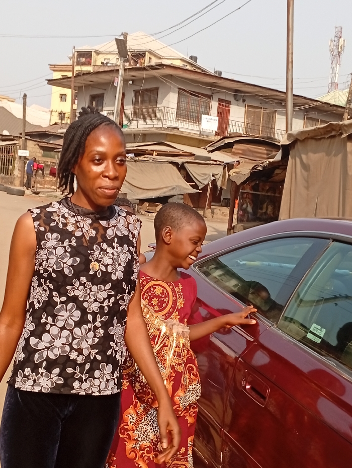 The image captures a vibrant daytime street scene, likely in an urban or semi-urban area, featuring two individuals – an adult woman and a young girl – alongside a car, with buildings and market stalls forming the background.
In the foreground on the left, an adult woman with dark skin and black braided hair, styled upwards, stands looking slightly to her left. She wears a sleeveless top with a black base and a prominent white and grey floral pattern. A gold-colored round pendant hangs from her neck. Her lower body is clad in dark, possibly navy blue or velvet-textured, trousers. Her expression is calm and neutral.
Beside her, on the right, stands a young girl with dark skin and very short, closely-cropped hair. She is wearing a striking sleeveless dress with a rich red or maroon background, adorned with elaborate gold, orange, and black patterns that include baroque swirls and possibly animal print elements. The girl is smiling broadly and appears joyful, looking towards the dark red/maroon sedan car parked beside her. Her right hand rests gently on the car's window frame. The car's window reflects distorted shapes from the surroundings.
The background reveals a bustling street environment. Immediately behind the individuals, on the left, are glimpses of what appear to be makeshift market stalls or small shops, covered with tarps and corrugated metal, suggesting commercial activity. Further back, a multi-story building dominates the upper part of the background. It is light-colored (likely white or light grey) with multiple windows, balconies, and railings, topped with a hip roof. On this building, a white rectangular sign with dark text is visible, which appears to read "TSASHEM CENTRE". To the far right, a utility pole or light pole extends upwards, and a communications tower is partially visible in the distance. The sky is clear and bright, indicating good weather. The overall impression is one of everyday life in a lively community.