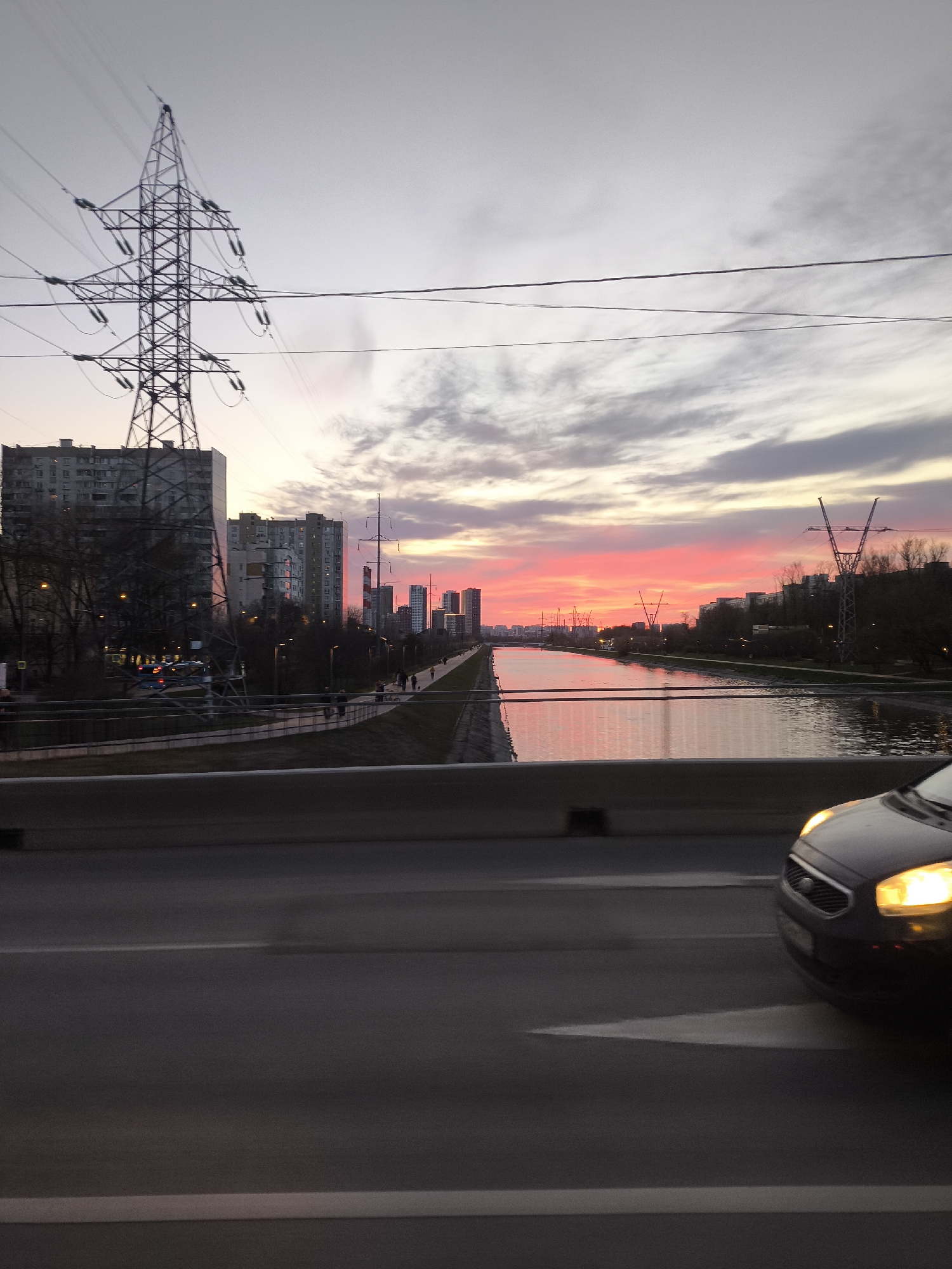The image is taken from a moving vehicle, likely a car, on a bridge over a canal in Moscow, Russia. A black car is partially visible in the foreground on the right side of the frame, with its headlights on. The road is dark, and white lane markings are visible.
In the midground, a canal reflects the colors of the sunset sky, primarily pink and orange. Concrete embankments line the canal, with a pedestrian walkway visible on the left bank. Several people are walking along the walkway.
In the background, high-rise buildings are silhouetted against the vibrant sunset. Power lines and electricity pylons are scattered throughout the scene, especially on the left side of the frame. The sky is a mix of grey, pink, and orange hues due to the sunset.
Overall, the image captures an urban scene with a canal, buildings, and infrastructure at sunset, likely in Moscow, Russia, with the shot taken from a moving car on a bridge.