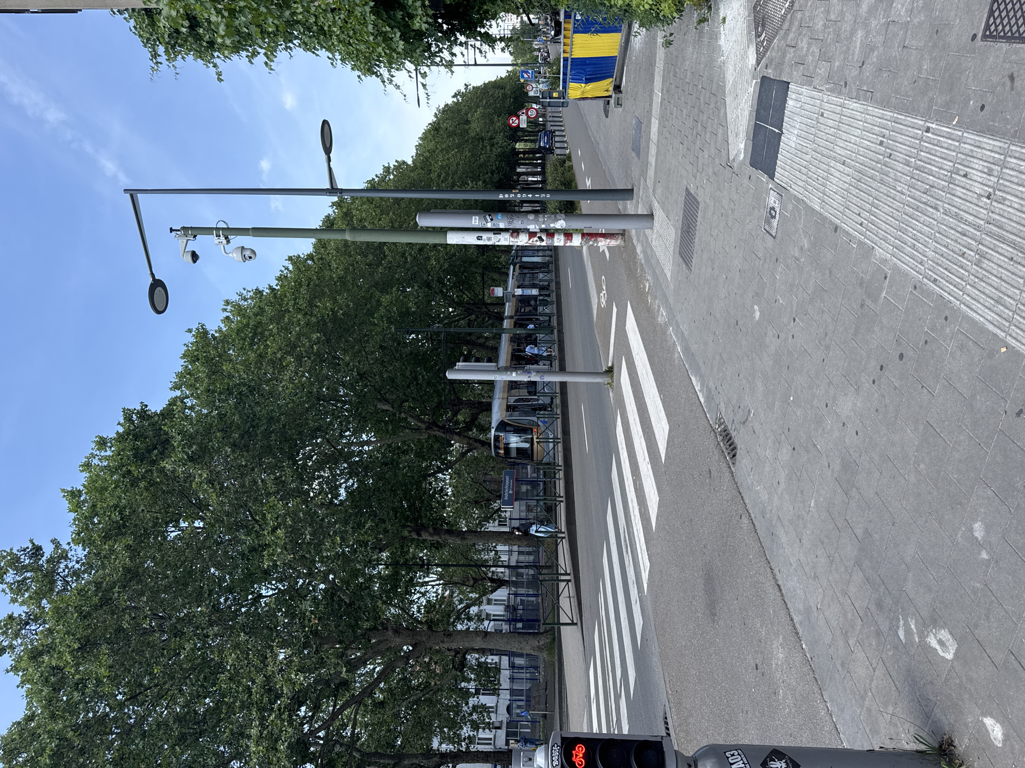 This is a vertical, eye-level shot of a street scene in Molenbeek-Saint-Jean, Belgium, likely taken during the daytime under clear skies. The foreground features a pedestrian crossing with bright white stripes painted on the asphalt. To the right of the crossing is a sidewalk paved with grey rectangular tiles, interspersed with textured tactile paving strips for the visually impaired. Several dark, square grates are embedded in the sidewalk.
A tall lamppost with two round, flat light fixtures stands near the sidewalk. Attached to another lamppost in the midground are two surveillance cameras, one white and dome-shaped, the other a more traditional white cylinder. This lamppost is also covered in various stickers. Beyond the lamppost, trees with dense green foliage line the street, casting dappled shadows.
In the background, a tram is visible at a stop. The tram is silver with dark windows and displays "49 Ans." on its side. A bus stop shelter with a dark roof and metal frame is also present. Further down the street, more trees and buildings can be seen. A yellow and blue construction barrier is also visible on the right side of the street.
A traffic light with a red bicycle symbol illuminated is in the bottom right corner of the image. A sticker with "COVI" is visible on the base of a lamppost in the foreground. The scene suggests a normal day with public transportation, pedestrian activity, and street maintenance.