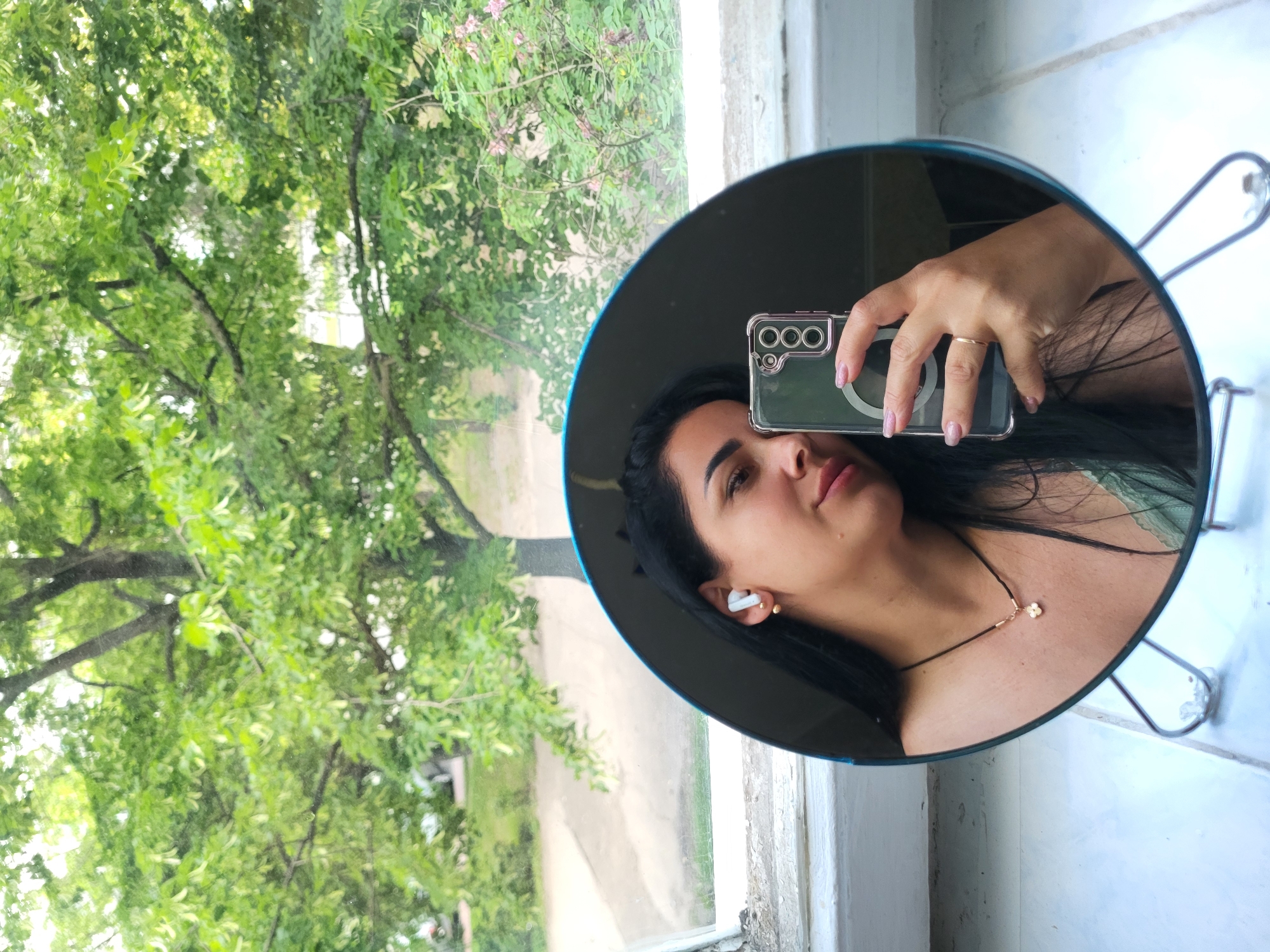 A young woman is taking a mirror selfie in front of a window. She is leaning to her right and her left ear is visible, with a white wireless earbud in it. She is wearing a necklace with a small clover charm. Her right hand is holding a smartphone with a clear case. The background shows lush green trees and vegetation, seen through the window. The mirror is round and supported by a metal stand. The scene appears to be indoors, near a window with a view of the outdoors. The location is not definitively identifiable as Horishni Plavni, Ukraine.